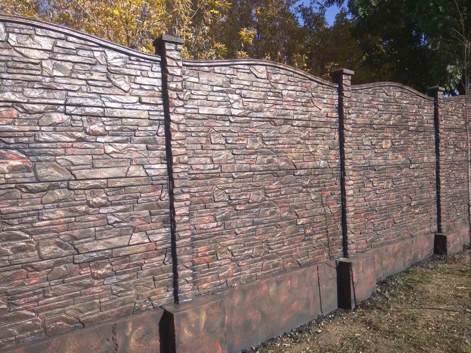 Dekoracyjny, betonowy płot imitujący mur z kamienia łupanego, z widocznymi filarami i falistym wykończeniem górnej krawędzi, w tle drzewa.