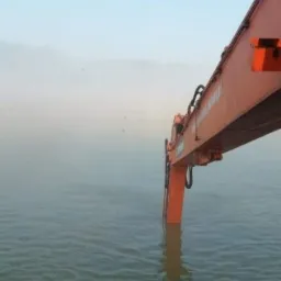 Pomarańczowe ramię koparki zanurzone w wodzie, widoczne z perspektywy brzegu, mglista atmosfera