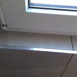 Bau-San - Fragment okna PCV z widocznym zawiasem, osadzonego w płytkach ceramicznych z metalową listwą wykończeniową.