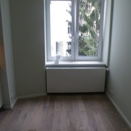 Bau-San - Pusty pokój z drewnianą podłogą, białym grzejnikiem pod oknem i fragmentem białej zabudowy kuchennej po lewej stronie. Przez okno widoczny budynek i drzewo.