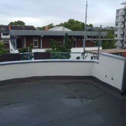 Bau-San - Płaski dach pokryty papą, częściowo mokry, z widocznymi detalami izolacji i ścianą attykową, w tle budynki mieszkalne i rusztowania.