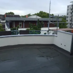 Płaski dach pokryty papą, częściowo mokry, z widocznymi detalami izolacji i ścianą attykową, w tle budynki mieszkalne i rusztowania.