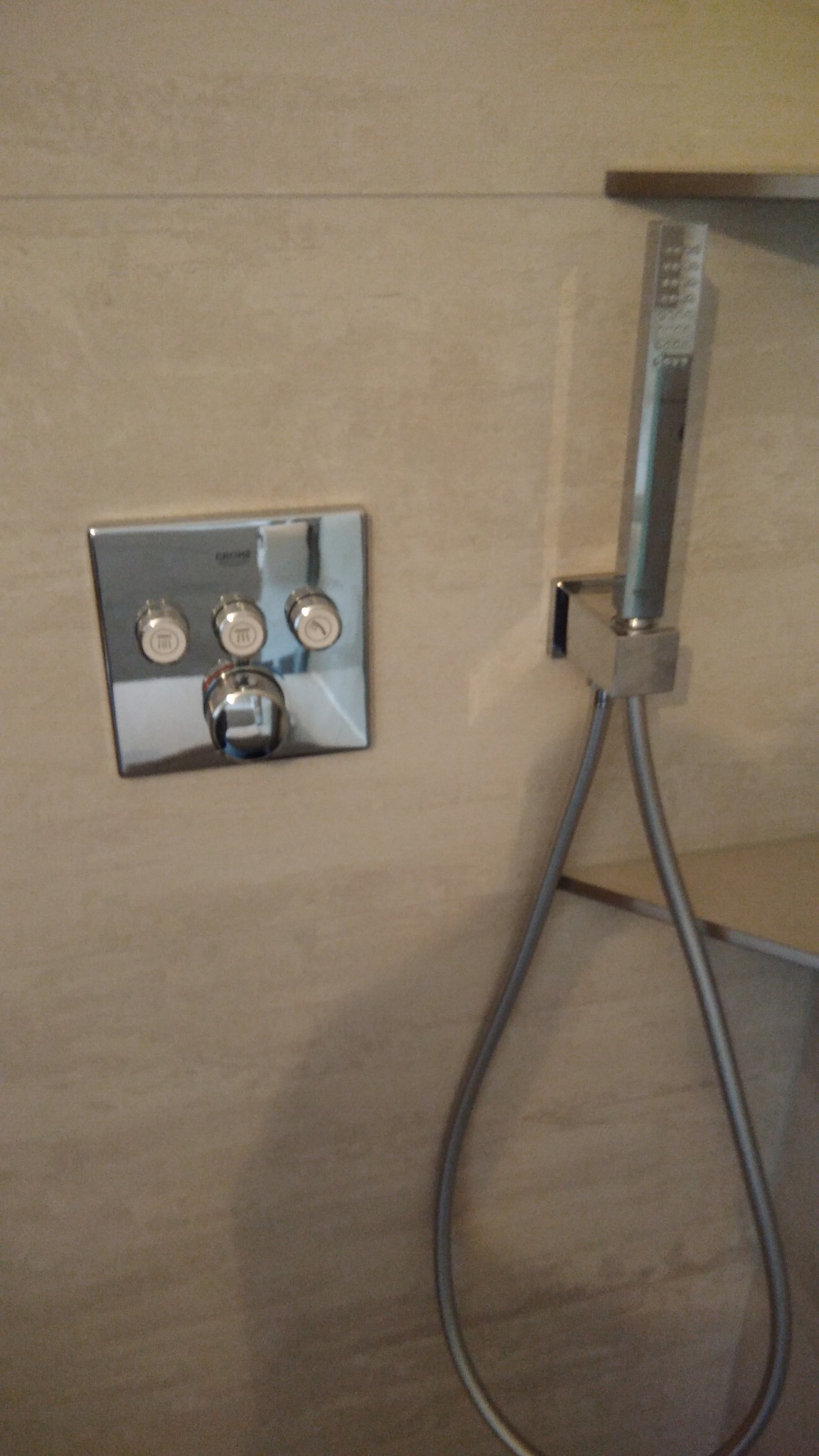 Chromowany panel sterowania prysznicem marki Grohe z trzema pokrętłami i wylewką, obok słuchawka prysznicowa na srebrnym wężu na tle beżowych płytek.