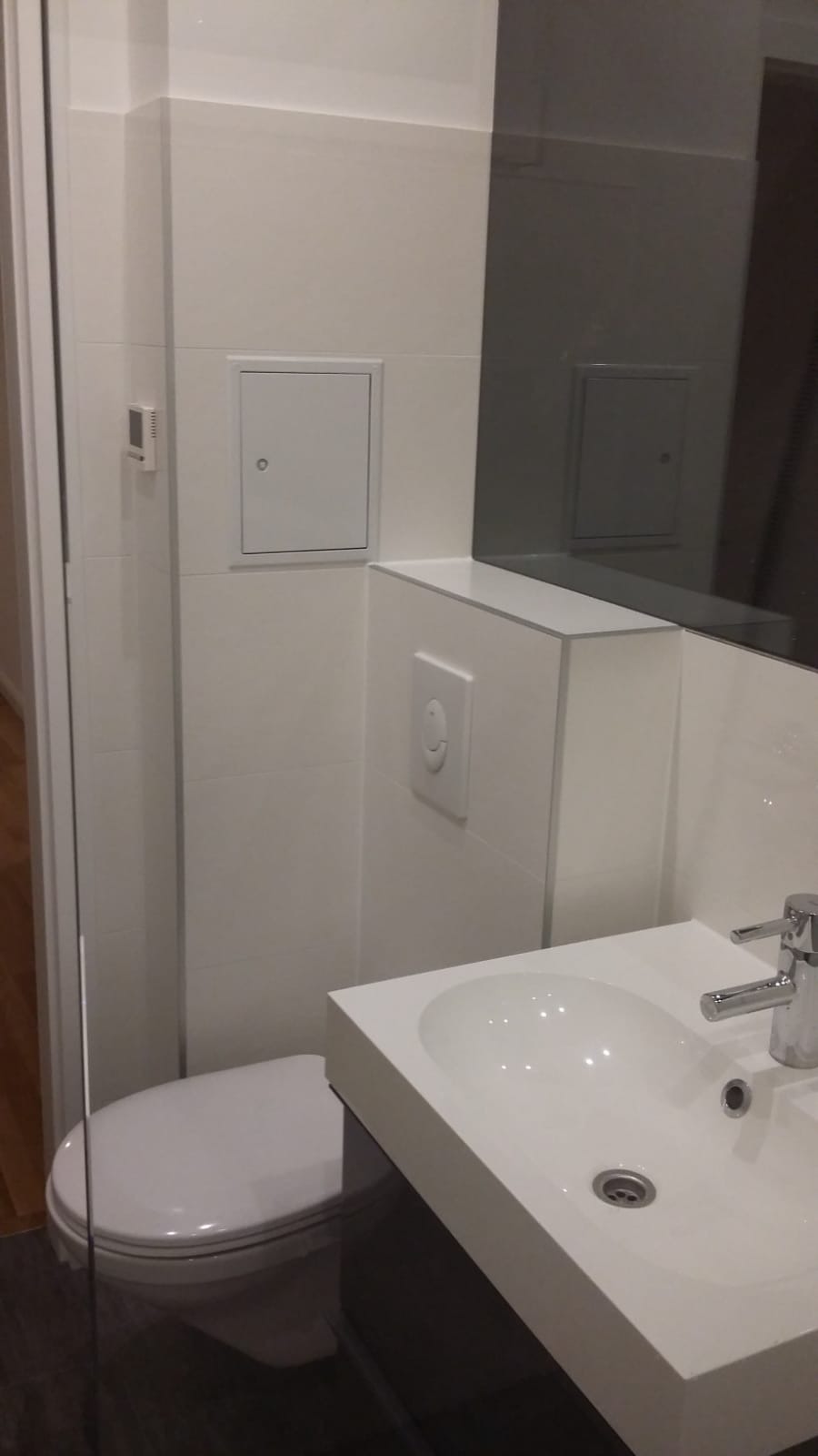 Nowoczesna, minimalistyczna łazienka z białymi płytkami, wiszącą toaletą, umywalką nablatową i dużym lustrem. Widoczny panel sterujący spłuczką i rewizja na ścianie.