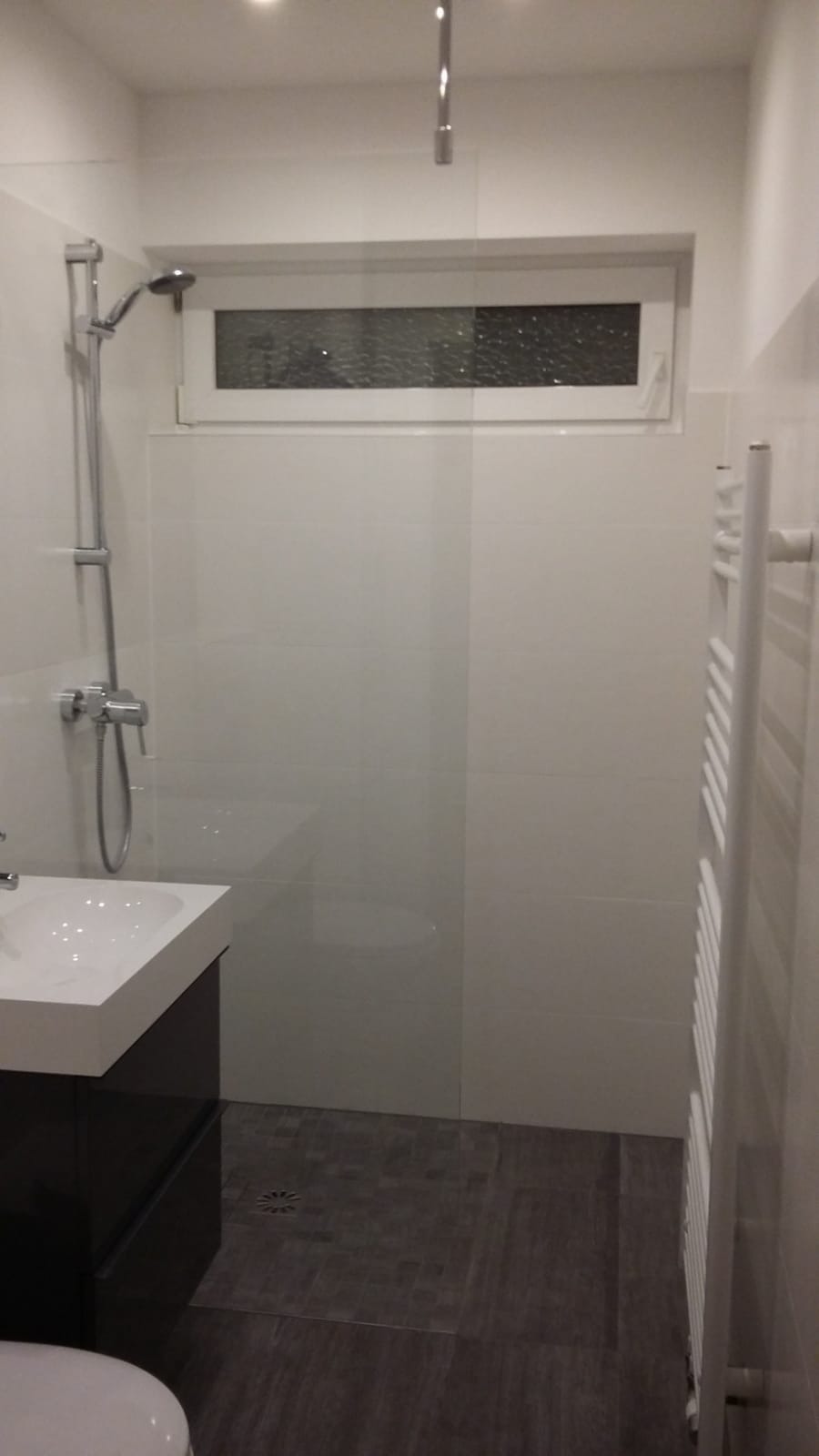 Nowoczesna łazienka z białymi płytkami, szarymi podłogowymi panelami, czarną szafką pod umywalką i grzejnikiem drabinkowym, oświetlona punktowo.