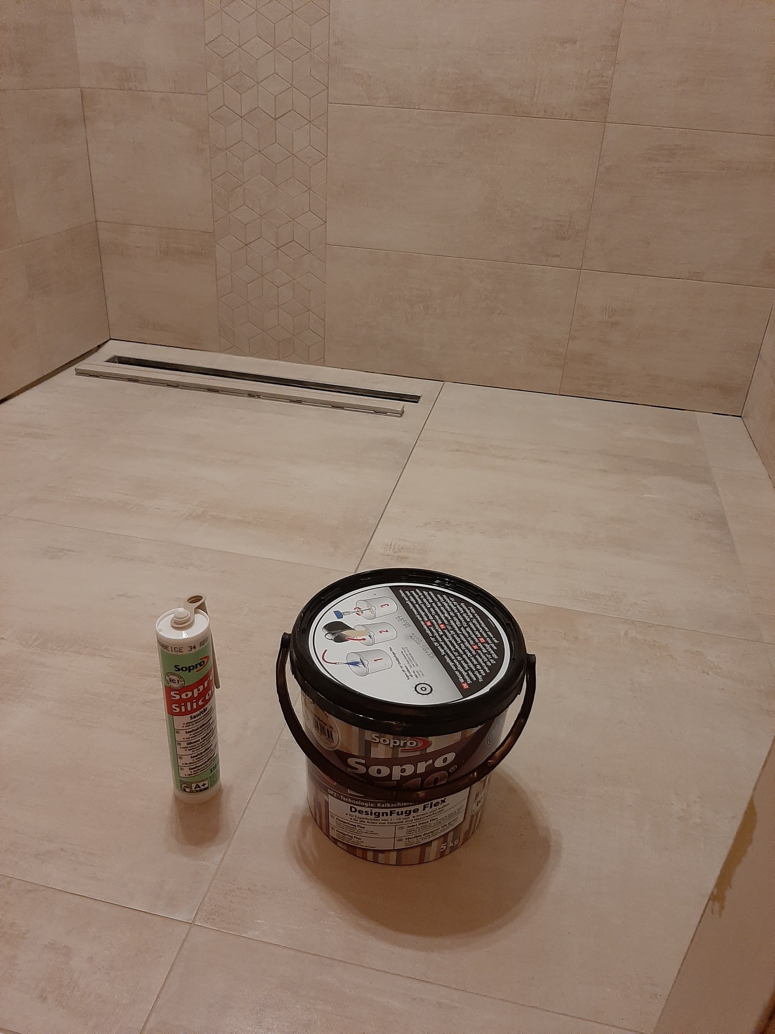 Przygotowania do fugowania w łazience: widoczny odpływ liniowy, płytki imitujące kamień z dekoracyjnym pasem mozaiki, wiadro z fugą Sopro DesignFuge Flex oraz tuba silikonu Sopro Sanitär.