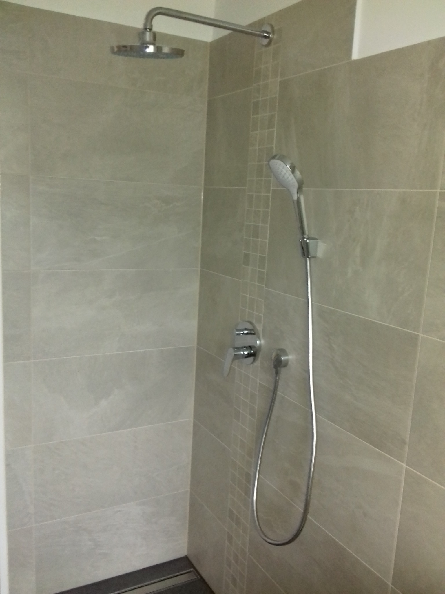 Szczegółowe ujęcie kabiny prysznicowej z szarymi płytkami, deszczownicą, słuchawką prysznicową na ścianie z dekoracyjnym paskiem mozaiki oraz widocznym odpływem liniowym.