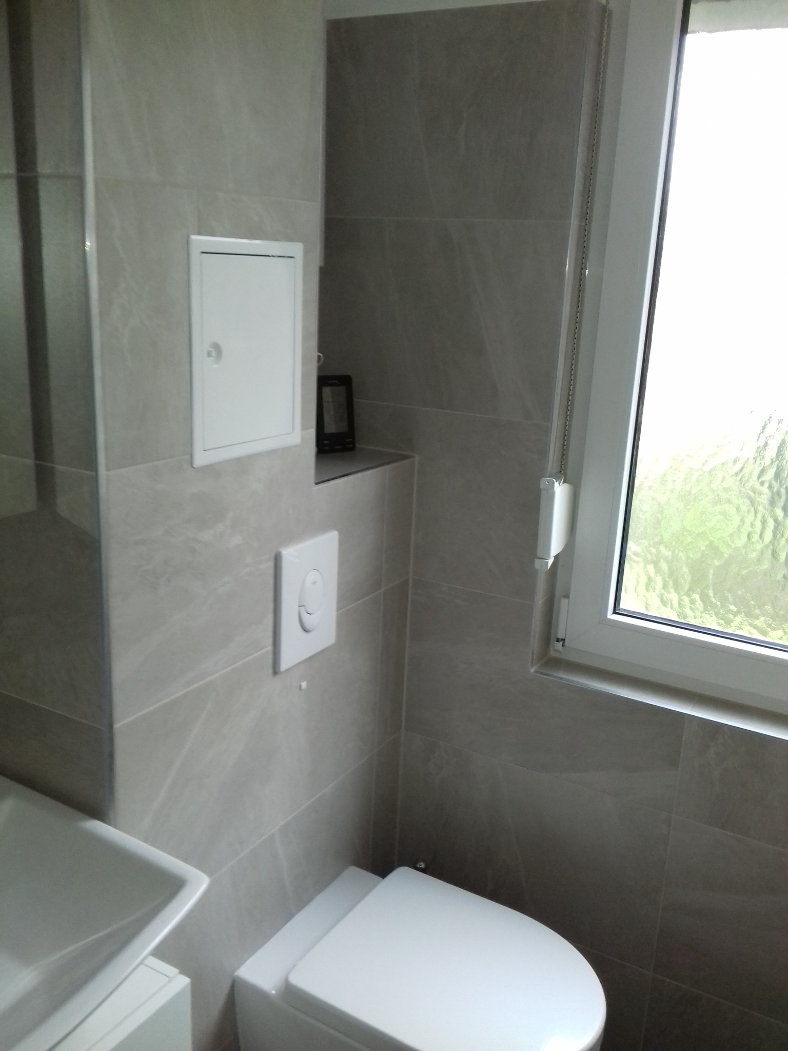 Nowoczesna toaleta z podtynkową spłuczką i ukrytą półką z czarnym czytnikiem e-booków, wykończona szarą, marmurkową płytką, oświetlona naturalnym światłem z okna z roletą.