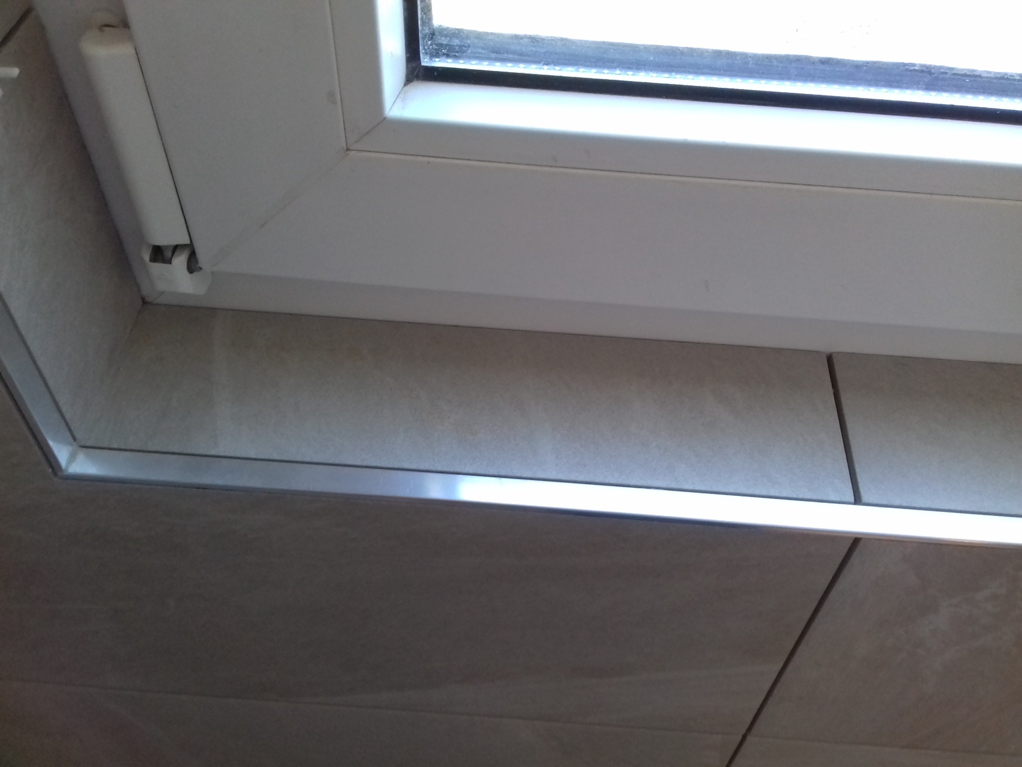 Fragment okna PCV z widocznym zawiasem, osadzonego w płytkach ceramicznych z metalową listwą wykończeniową.