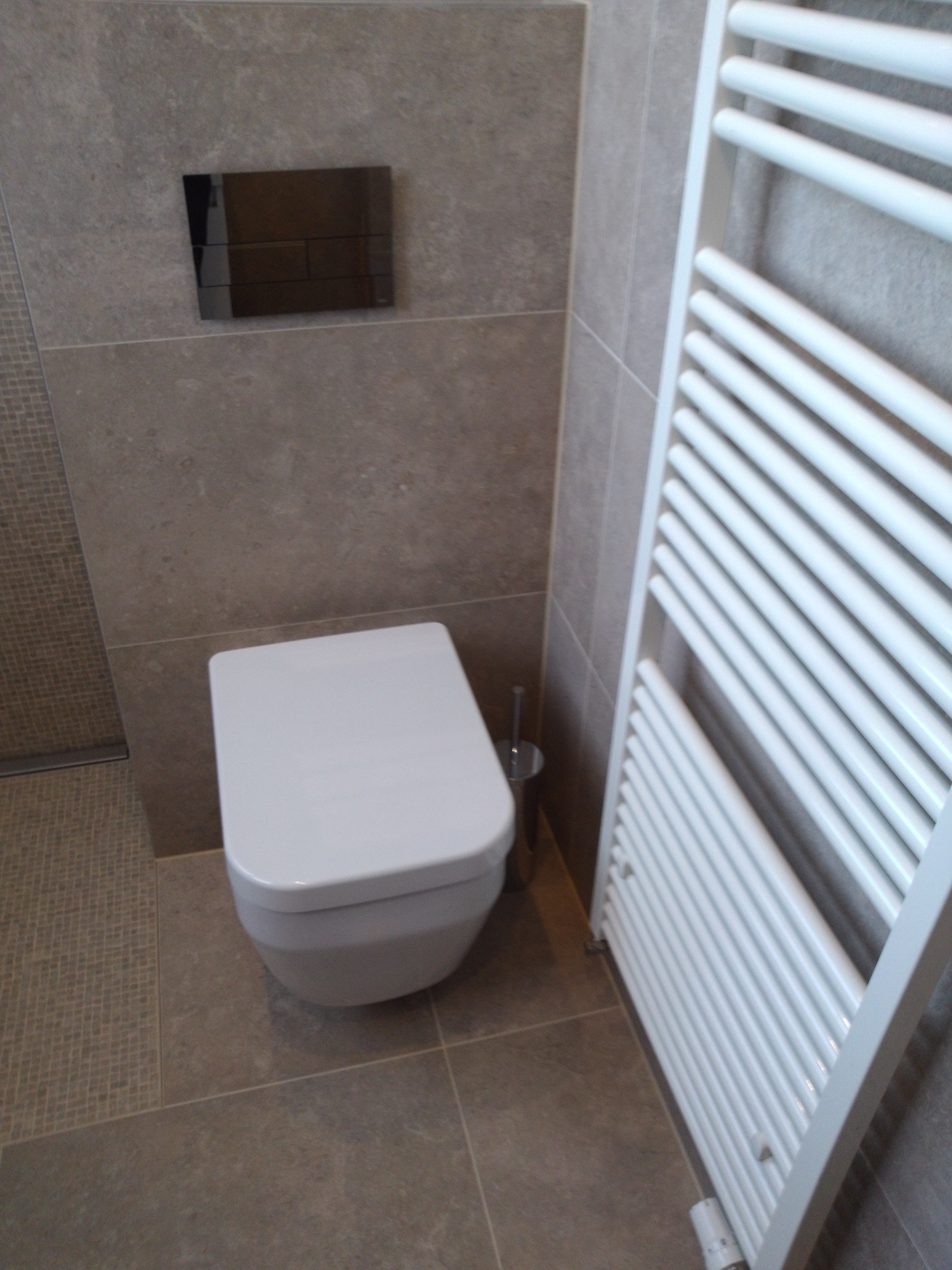 Nowoczesna toaleta podwieszana z czarnym przyciskiem spłukującym na szarej, gładkiej ścianie, obok biały grzejnik drabinkowy i szczotka do WC.