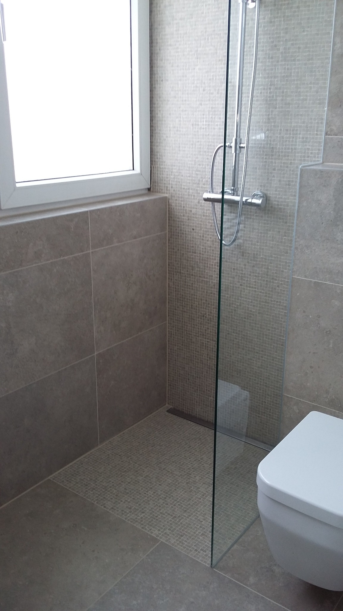 Nowoczesna kabina prysznicowa ze szklaną ścianką, wyłożona mozaiką, z widocznym odpływem liniowym i podwieszaną toaletą w minimalistycznej łazience z oknem.