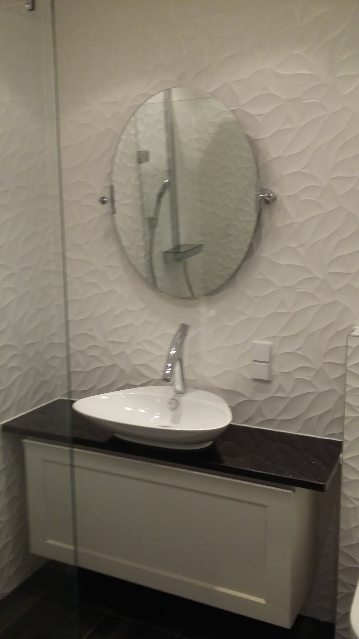 Nowoczesna umywalka nablatowa o owalnym kształcie z chromowaną baterią, zamontowana na szafce z ciemnym blatem, na tle ściany z dekoracyjnymi, falistymi płytkami i okrągłym lustrem.