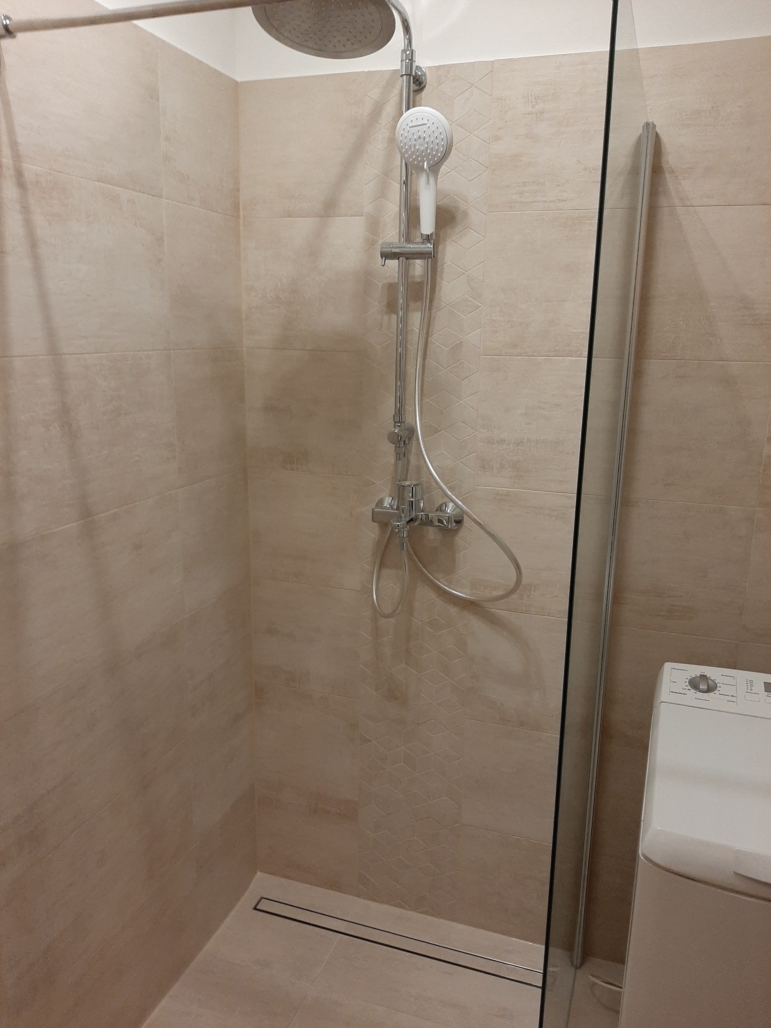 Beżowa kabina prysznicowa z deszczownicą, panelem prysznicowym z białą słuchawką i odpływem liniowym, przylegająca do pralki.