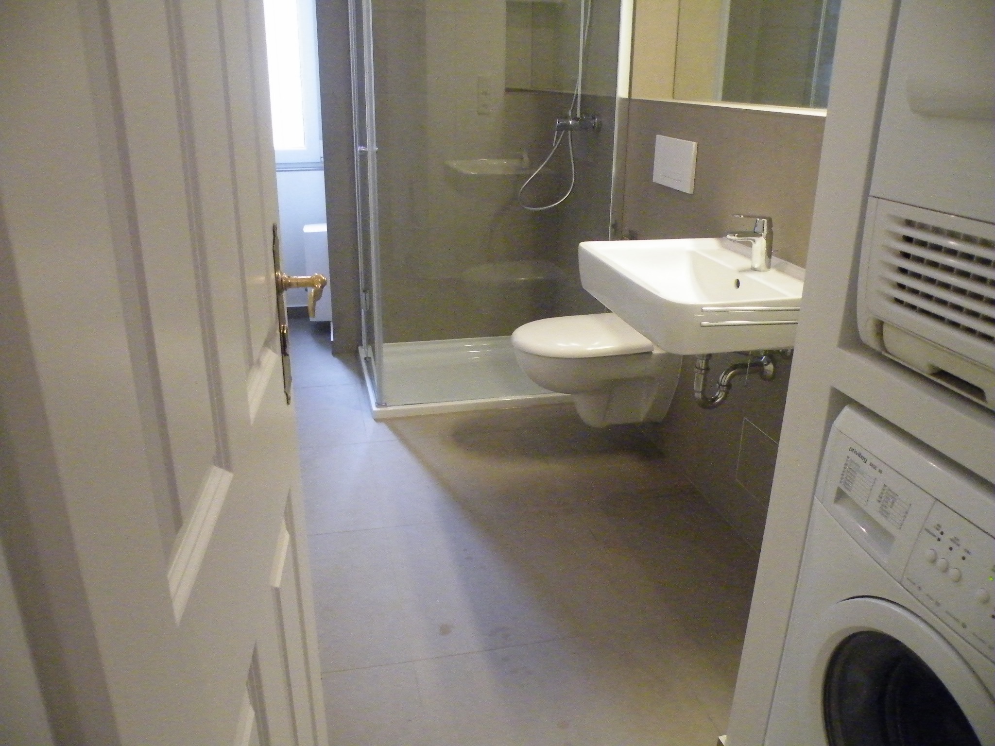 Otwarta biała łazienka z kabiną prysznicową, umywalką wiszącą, toaletą i pralką, widok zza częściowo otwartych drzwi.