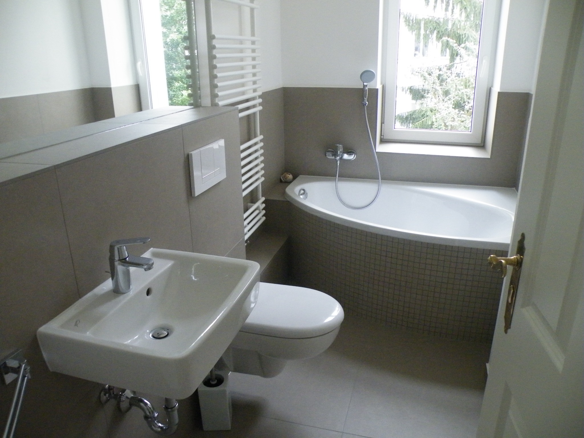 Jasna łazienka z narożną wanną obłożoną mozaiką, umywalką nablatową z chromowaną baterią, wiszącą toaletą i grzejnikiem drabinkowym przy oknie.