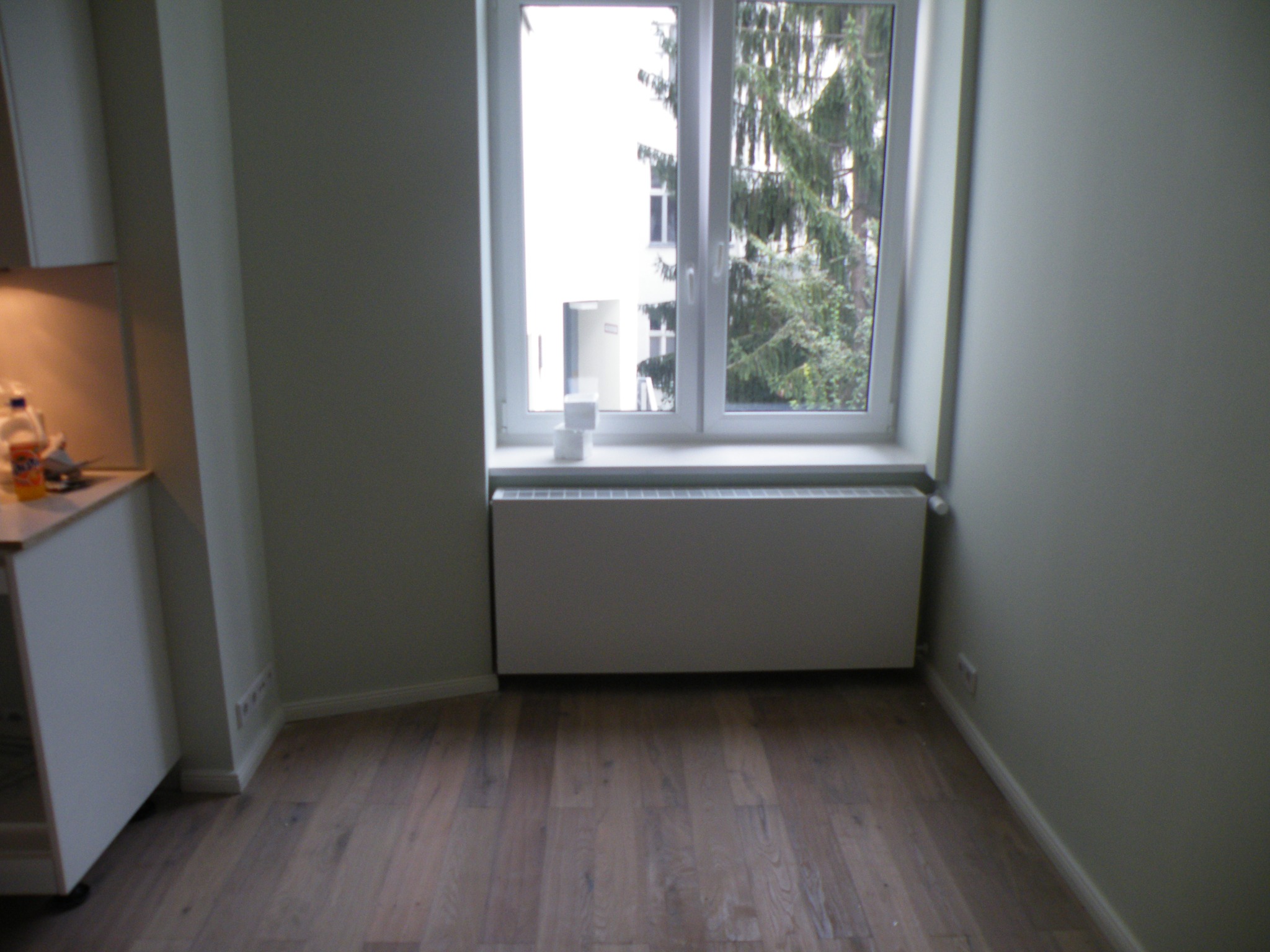 Pusty pokój z drewnianą podłogą, białym grzejnikiem pod oknem i fragmentem białej zabudowy kuchennej po lewej stronie. Przez okno widoczny budynek i drzewo.