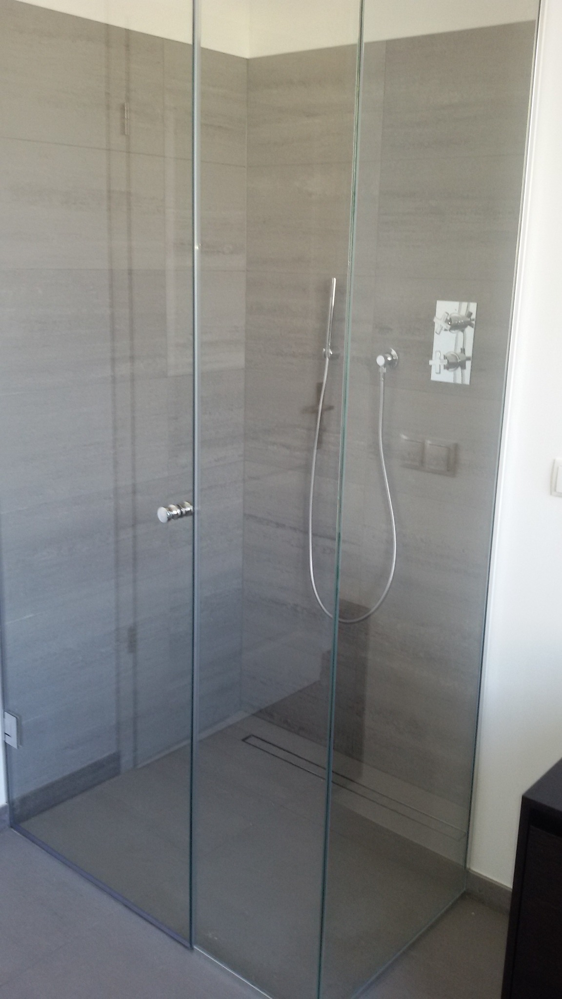 Szklana kabina prysznicowa typu walk-in z odpływem liniowym w podłodze, wykończona szarymi płytkami imitującymi kamień.