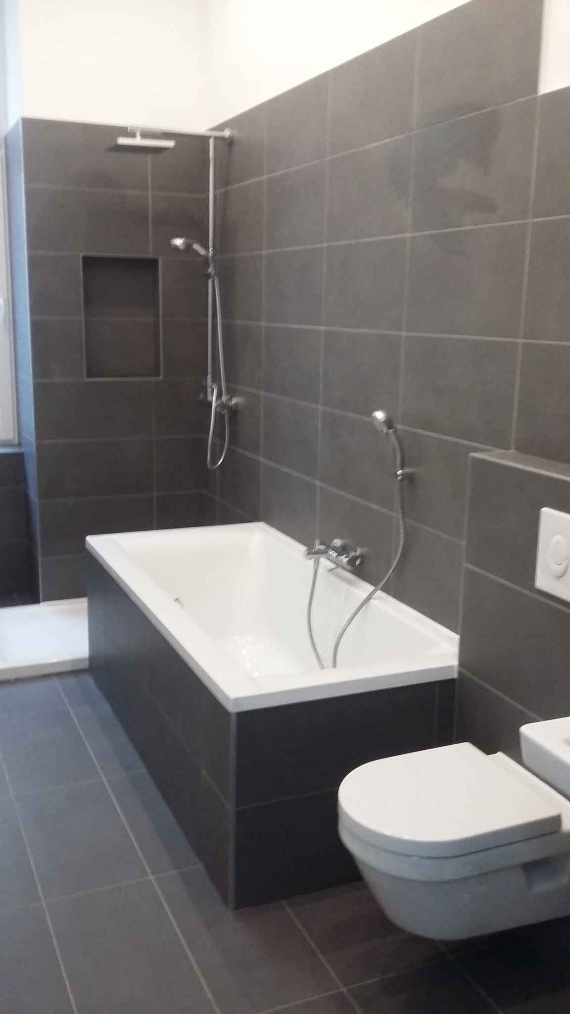 Nowoczesna łazienka w odcieniach szarości z prostokątną wanną, kabiną prysznicową typu walk-in i podwieszaną toaletą.