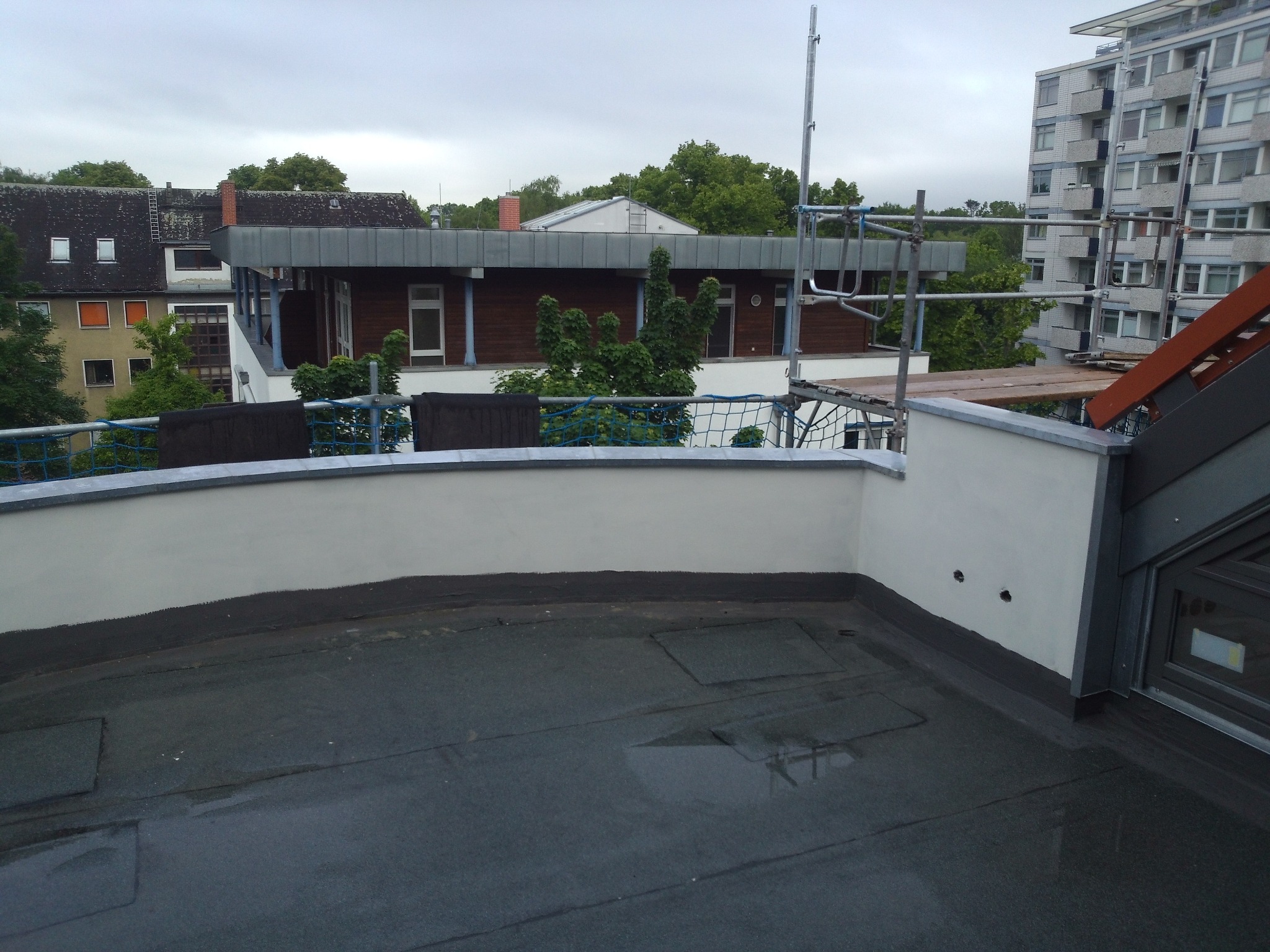 Płaski dach pokryty papą, częściowo mokry, z widocznymi detalami izolacji i ścianą attykową, w tle budynki mieszkalne i rusztowania.