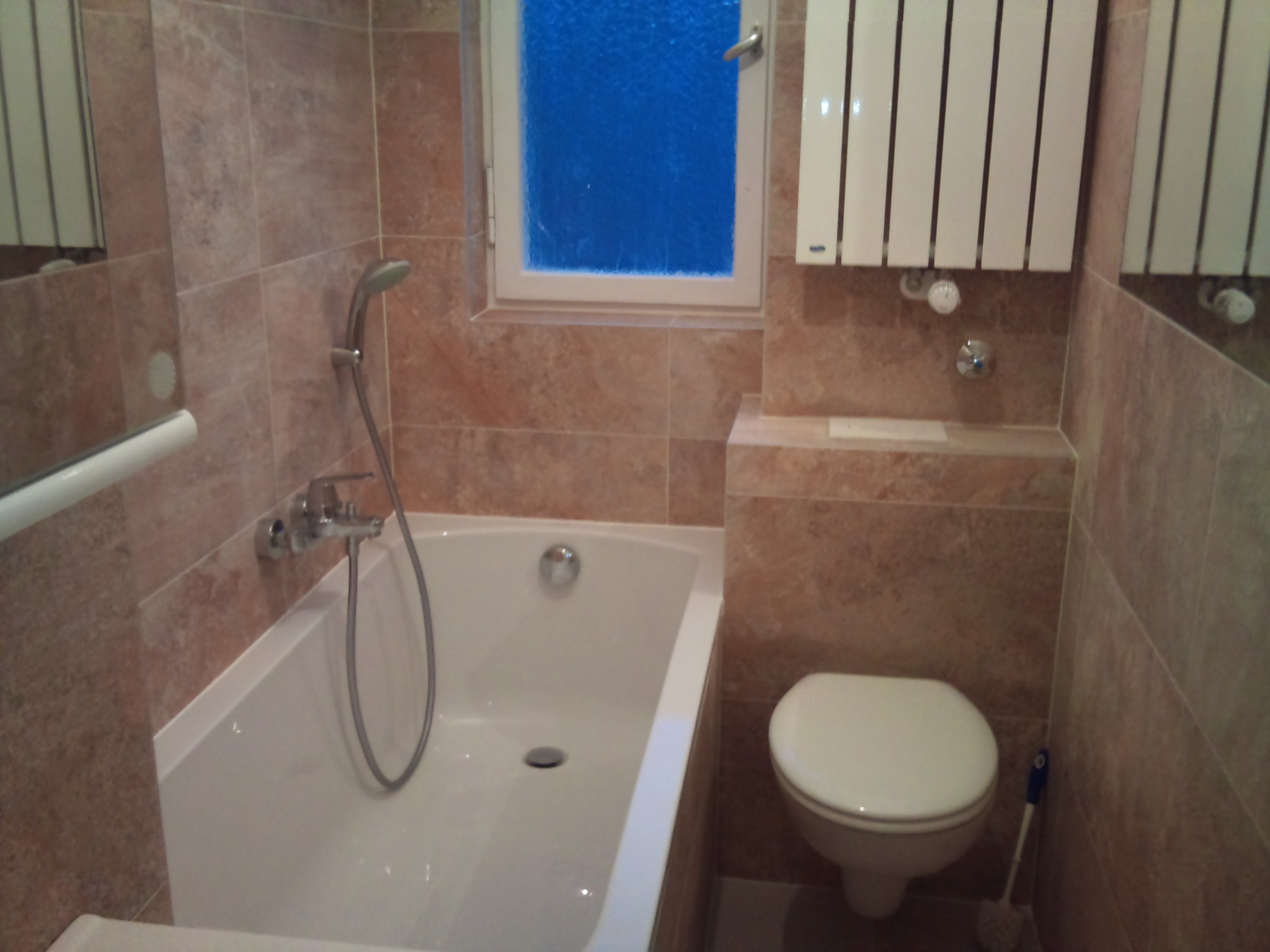 Wykończona łazienka z beżowymi płytkami, białą wanną, toaletą i grzejnikiem żeberkowym pod oknem z niebieską szybą.