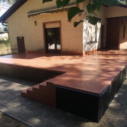 Mateusz Hapke - Wykończony taras z żywicy epoksydowej przy domu jednorodzinnym. Widoczne schody i fragment elewacji z oknami. Całość utrzymana w ciepłych, brązowych tonacjach.