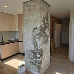 Mateusz Hapke - Kolumna z dekoracyjną fakturą i motywem kwiatowym w jasnym wnętrzu z drewnianą podłogą i zabudową kuchenną. Widoczne narzędzia i materiały budowlane.