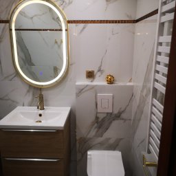 Mateusz Hapke - Elegancka łazienka z marmurowymi płytkami, podświetlanym lustrem, drewnianą szafką pod umywalką i grzejnikiem drabinkowym. Wykończenie w odcieniach bieli, złota i brązu.