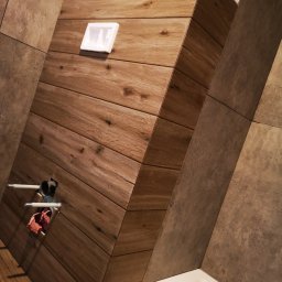 Mateusz Hapke - Nowoczesna zabudowa WC z drewnopodobnych płytek w łazience, widoczne przyłącza wodne i fragment wanny. Nietypowy układ płytek na ścianie.