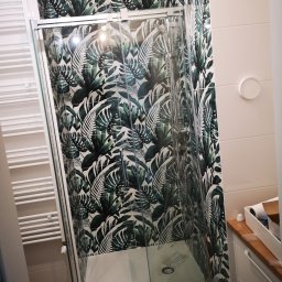 Mateusz Hapke - Nowoczesna kabina prysznicowa z deszczownicą, otoczona płytkami w tropikalne liście. Biały grzejnik drabinkowy i drewniany blat dodają ciepła aranżacji.