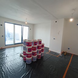 Mateusz Hapke - Przygotowania do malowania: pokój zabezpieczony folią, ustawione wiadra z farbą Atlas Rapid. Widoczne gładkie ściany, okno z niebieską taśmą malarską i narzędzia.