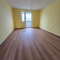 Mateusz Hapke - Pusty pokój z panelami podłogowymi w odcieniach brązu i żółtymi ścianami. Widoczne okno balkonowe i sufit z wiszącym przewodem. Podłoga ułożona wzdłuż dłuższego boku pokoju.