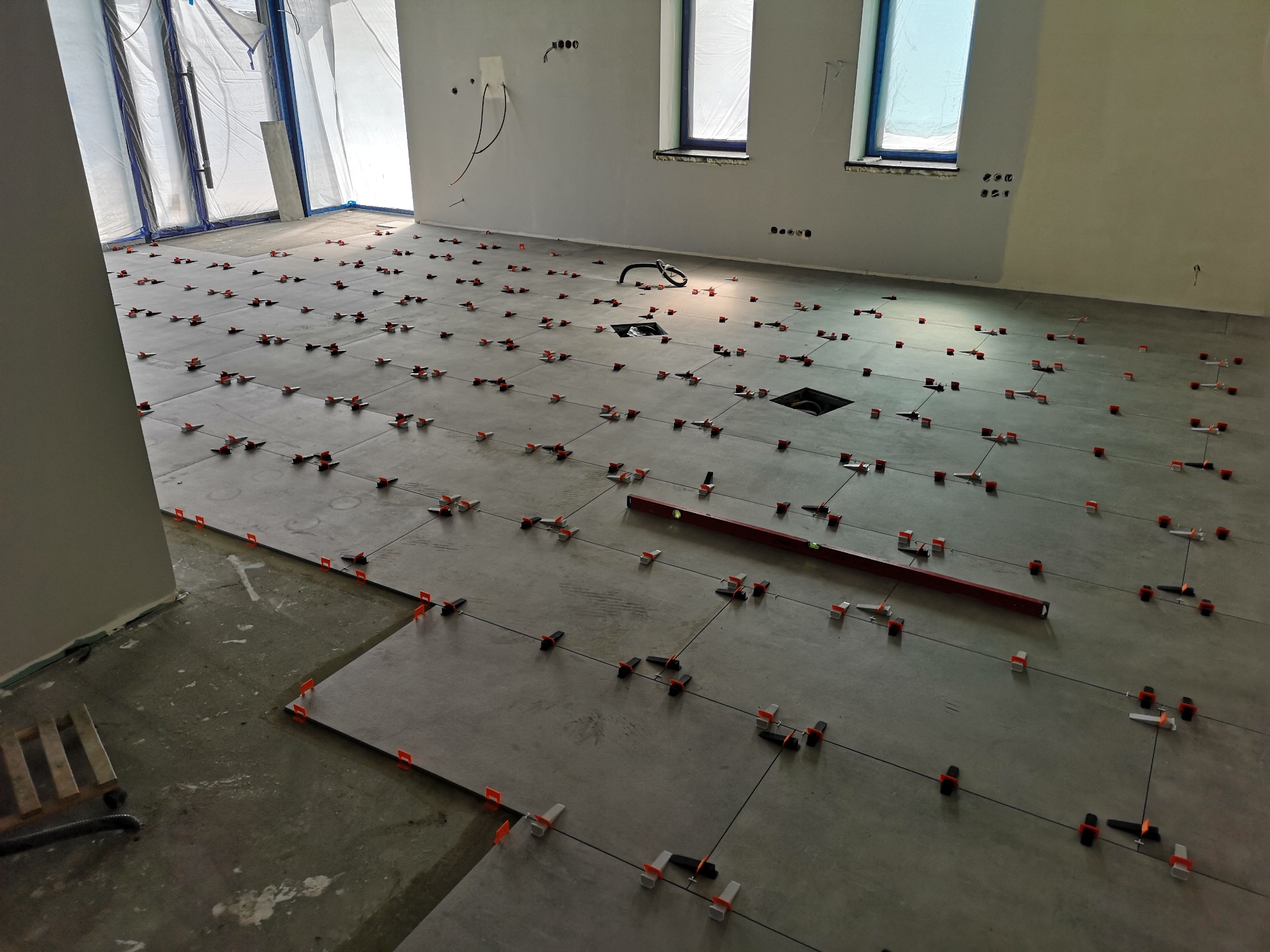 Szare płytki podłogowe w trakcie układania, z systemem poziomowania płytek, widoczna poziomica, okna zabezpieczone folią malarską, przygotowane otwory w podłodze.