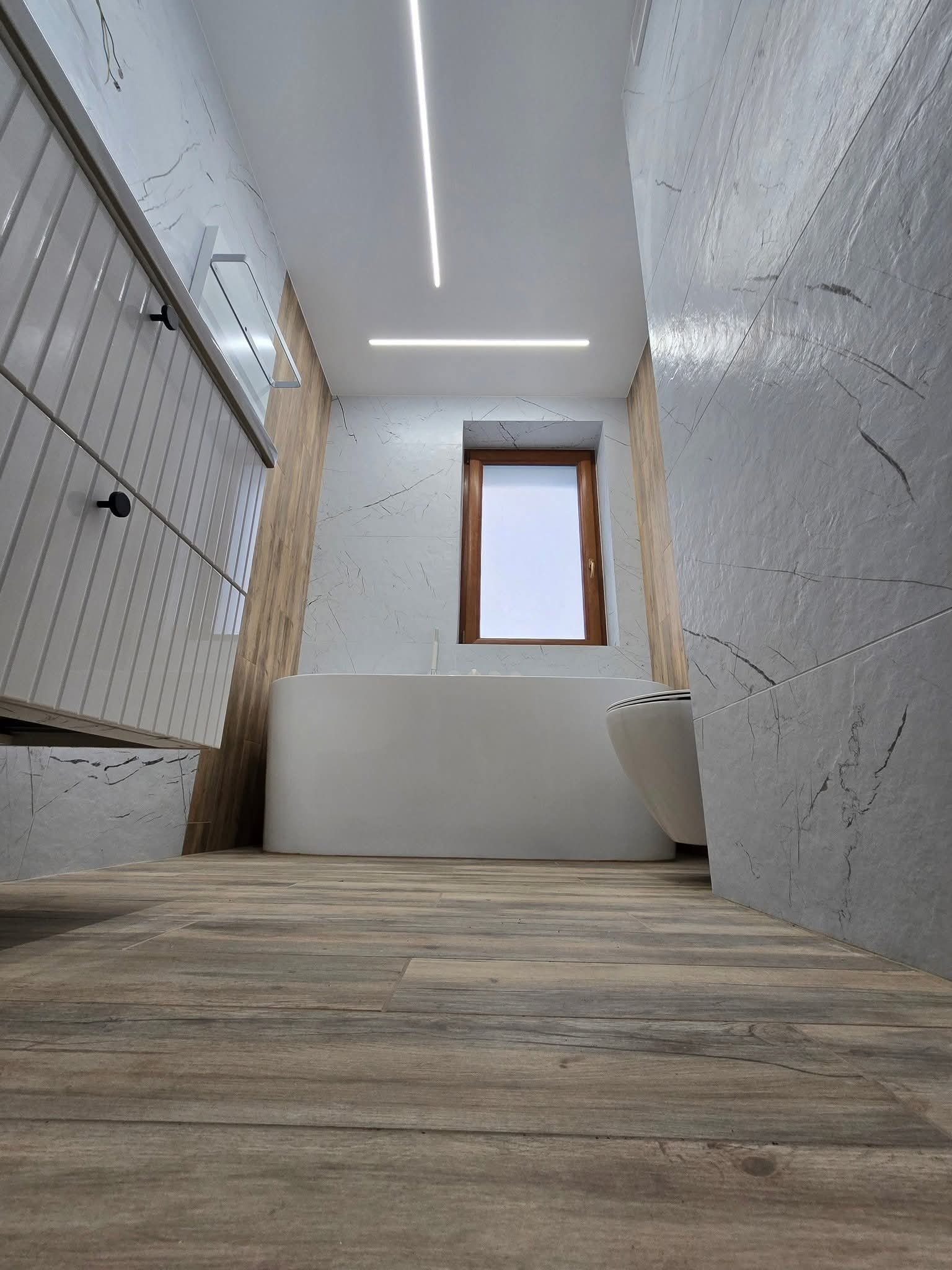 Nowoczesna łazienka w Poznaniu z drewnopodobną podłogą, białą wanną i umywalką. Ściany wykończone płytkami imitującymi marmur. Minimalistyczne oświetlenie.