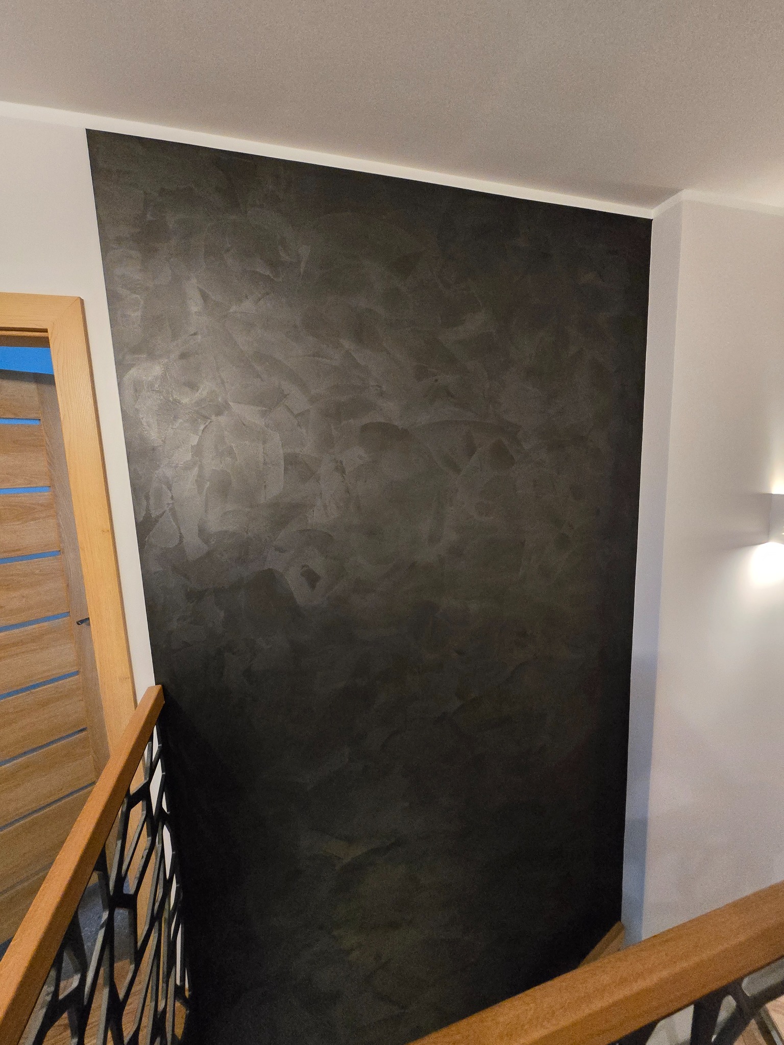 Ściana w ciemnym odcieniu z efektem dekoracyjnym, widoczna z perspektywy schodów. Fragment drewnianej balustrady i drzwi w tle. Oświetlenie punktowe.