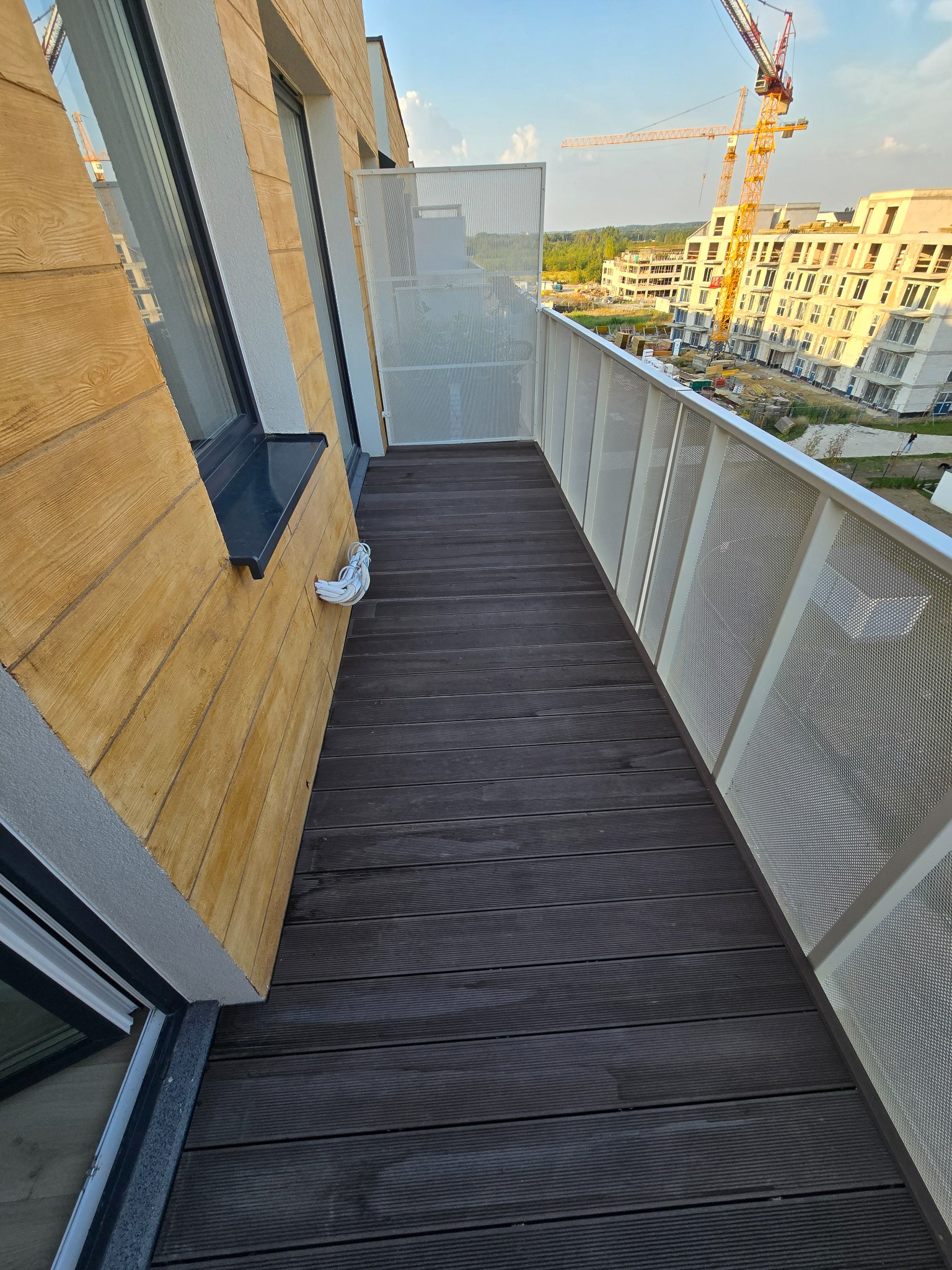 Nowoczesny balkon z drewnianą podłogą i ażurową balustradą. Widok na budowę z żurawiami w tle, jasne drewno elewacji i okno z czarną ramą.