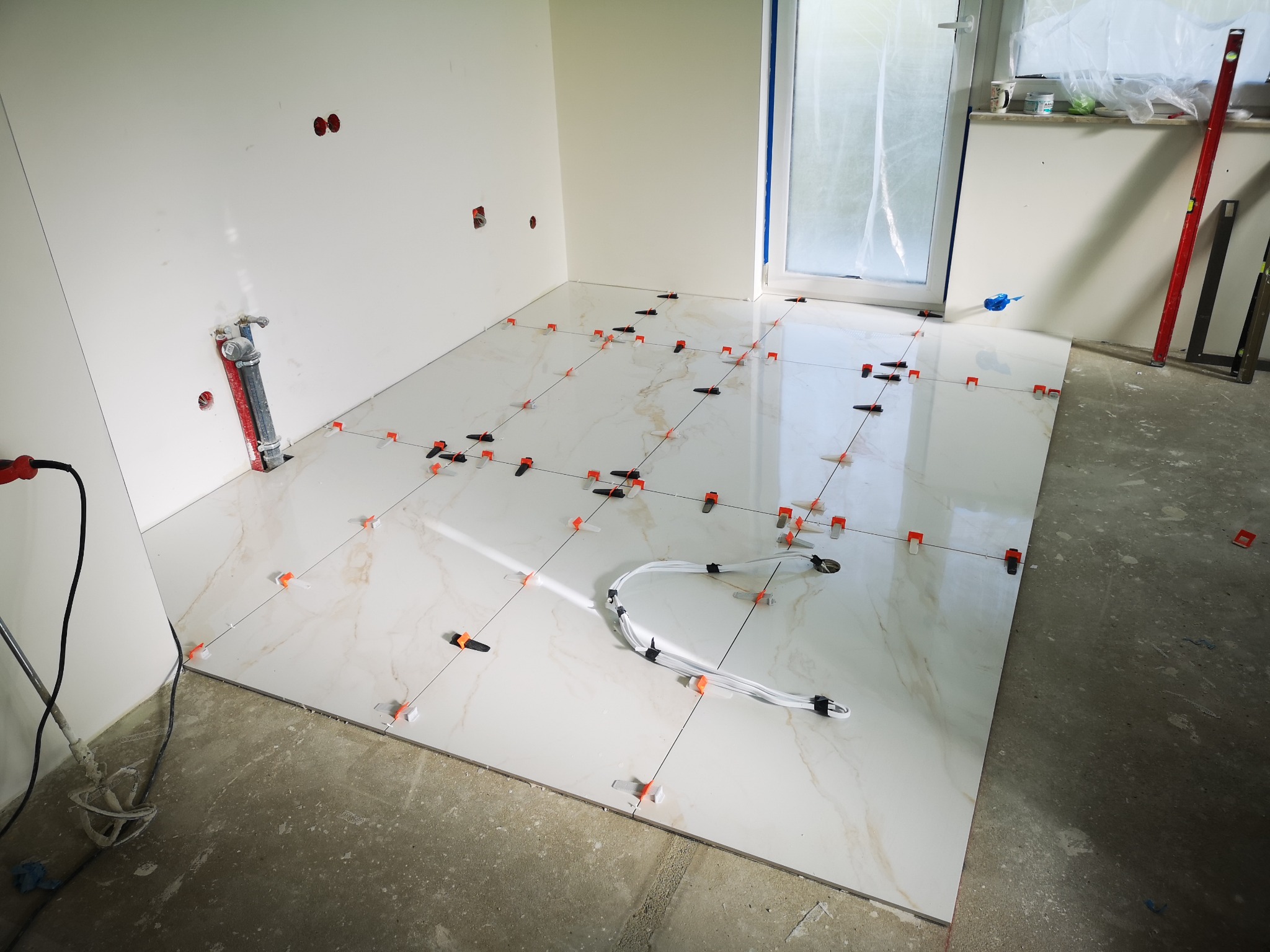 Układanie płytek podłogowych w jasnym wnętrzu z użyciem klipsów poziomujących, widoczne przewody i narzędzia. Etap prac glazurniczych w trakcie realizacji.