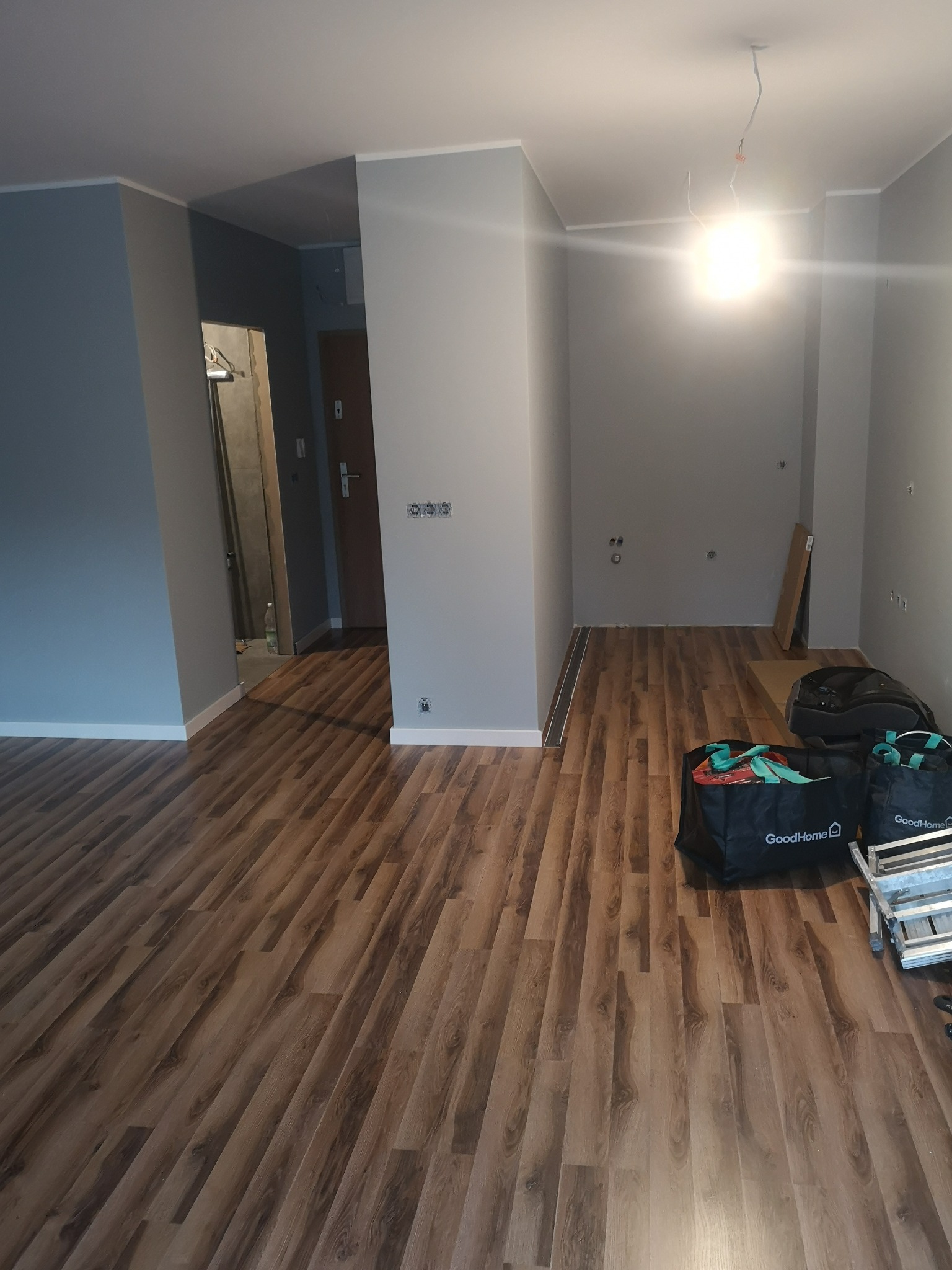 Szare ściany i drewnopodobna podłoga w pustym pokoju po remoncie. Widoczne torby z napisem GoodHome i drabina. Przygotowanie do dalszych prac wykończeniowych.