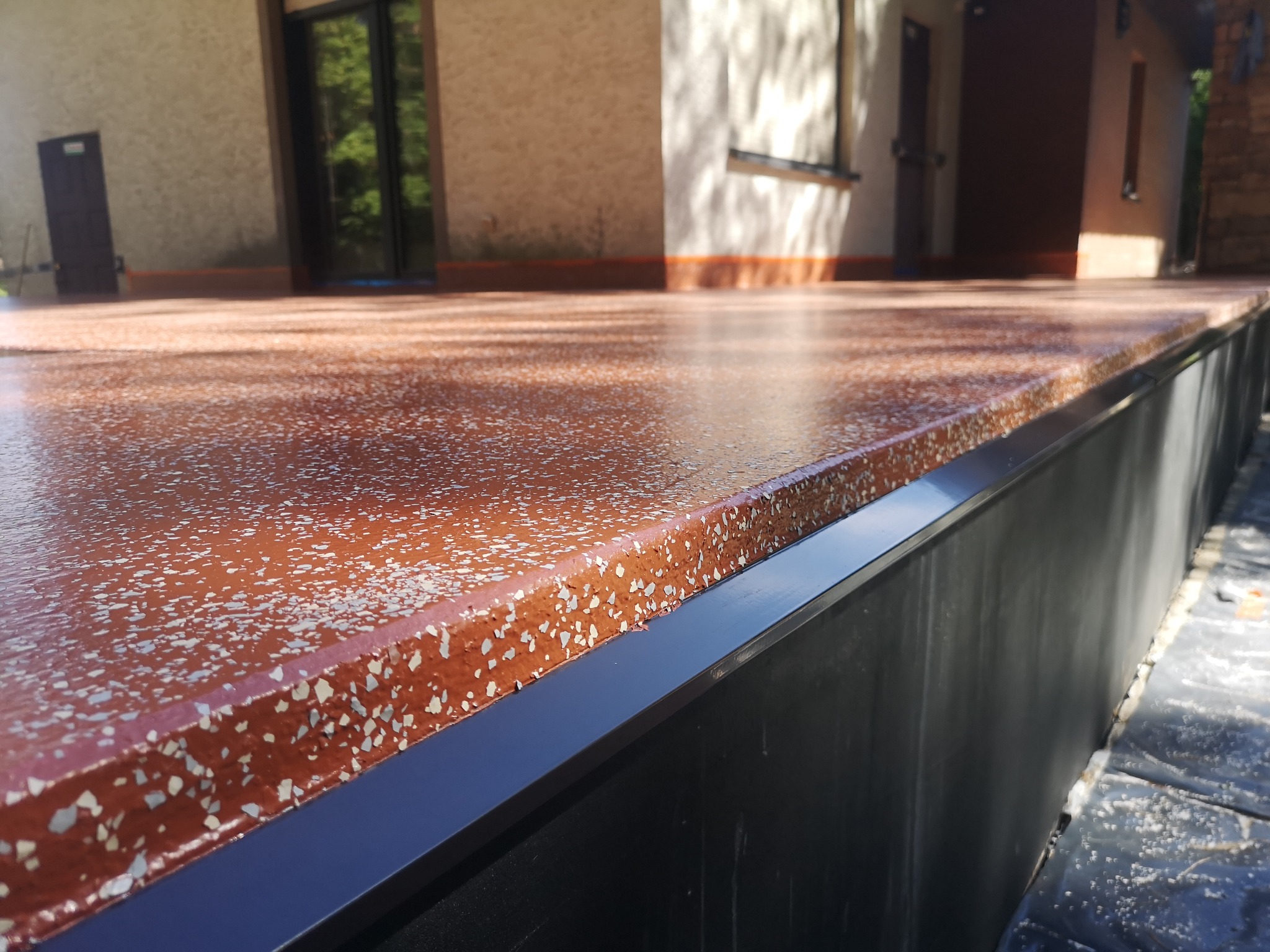 Błyszcząca, czerwono-brązowa posadzka tarasu z białymi inkluzjami, wykończona czarną listwą. Widok z bliska na krawędź i elewację budynku w tle.