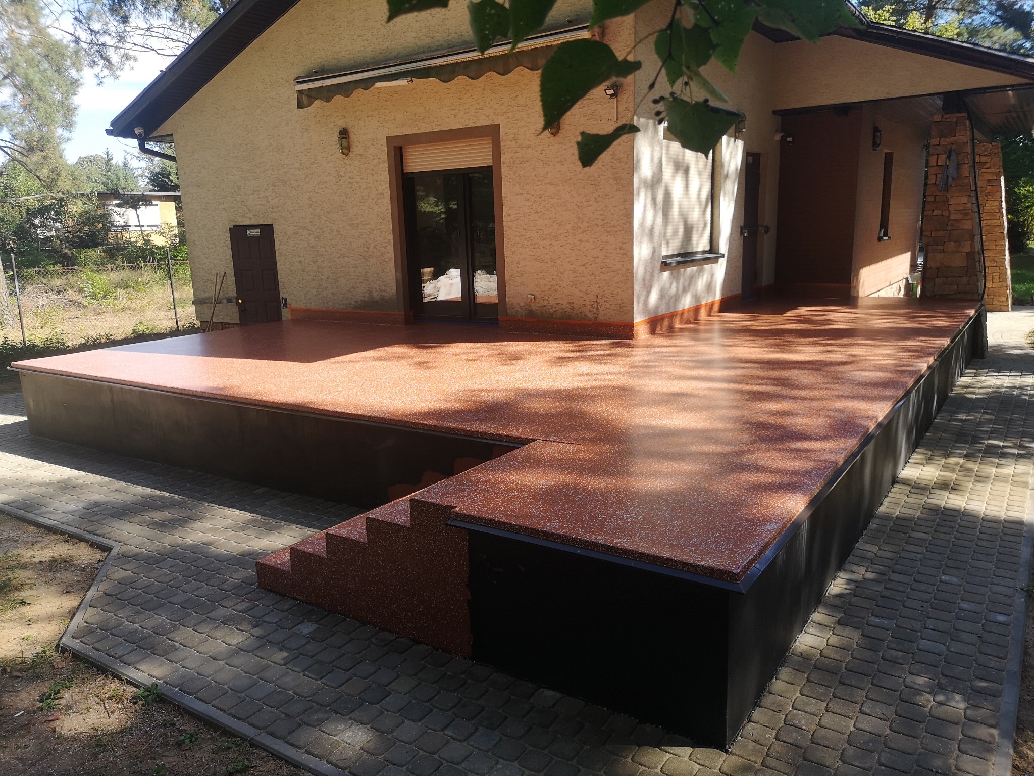 Wykończony taras z żywicy epoksydowej przy domu jednorodzinnym. Widoczne schody i fragment elewacji z oknami. Całość utrzymana w ciepłych, brązowych tonacjach.