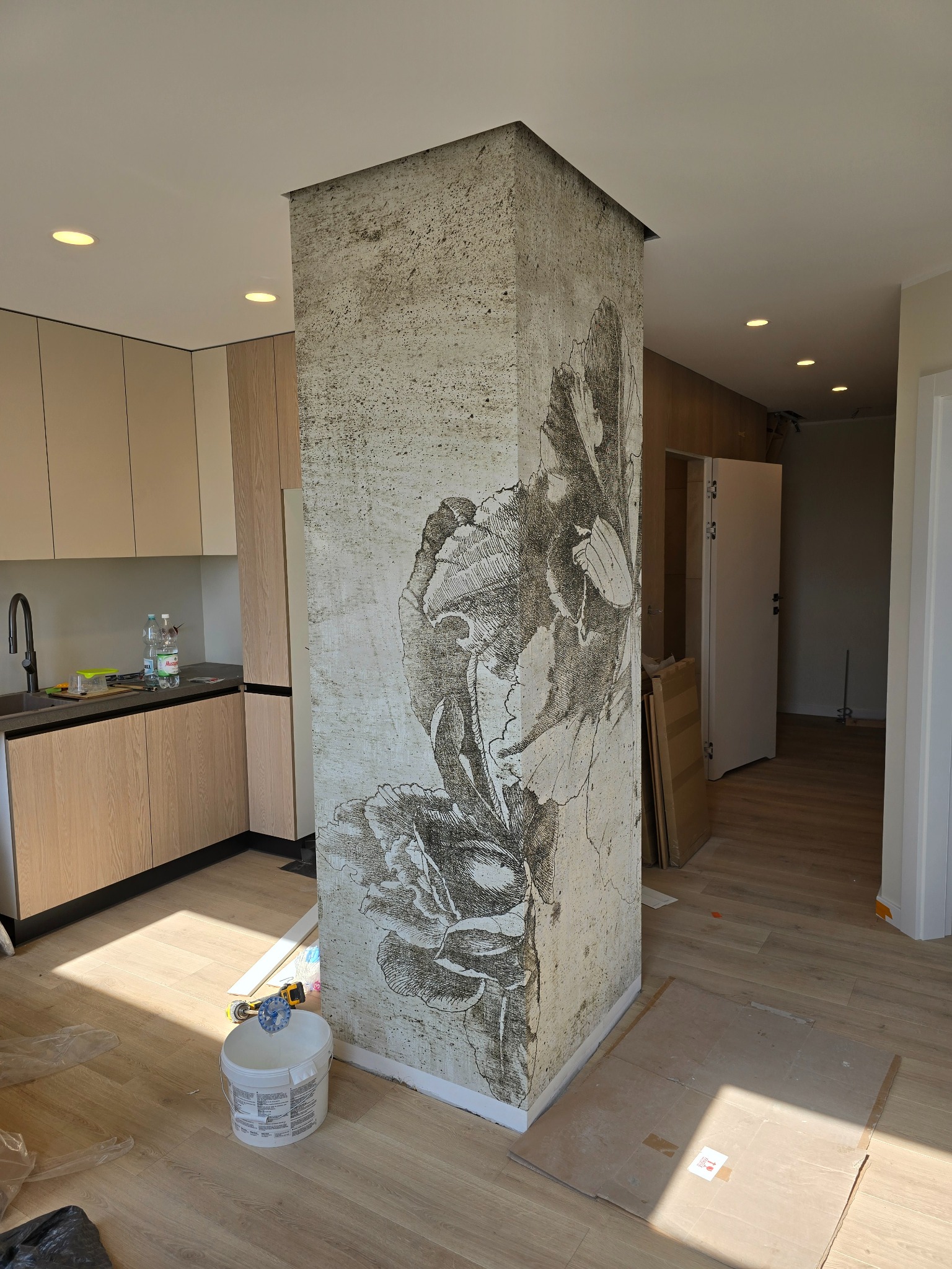 Kolumna z dekoracyjną fakturą i motywem kwiatowym w jasnym wnętrzu z drewnianą podłogą i zabudową kuchenną. Widoczne narzędzia i materiały budowlane.