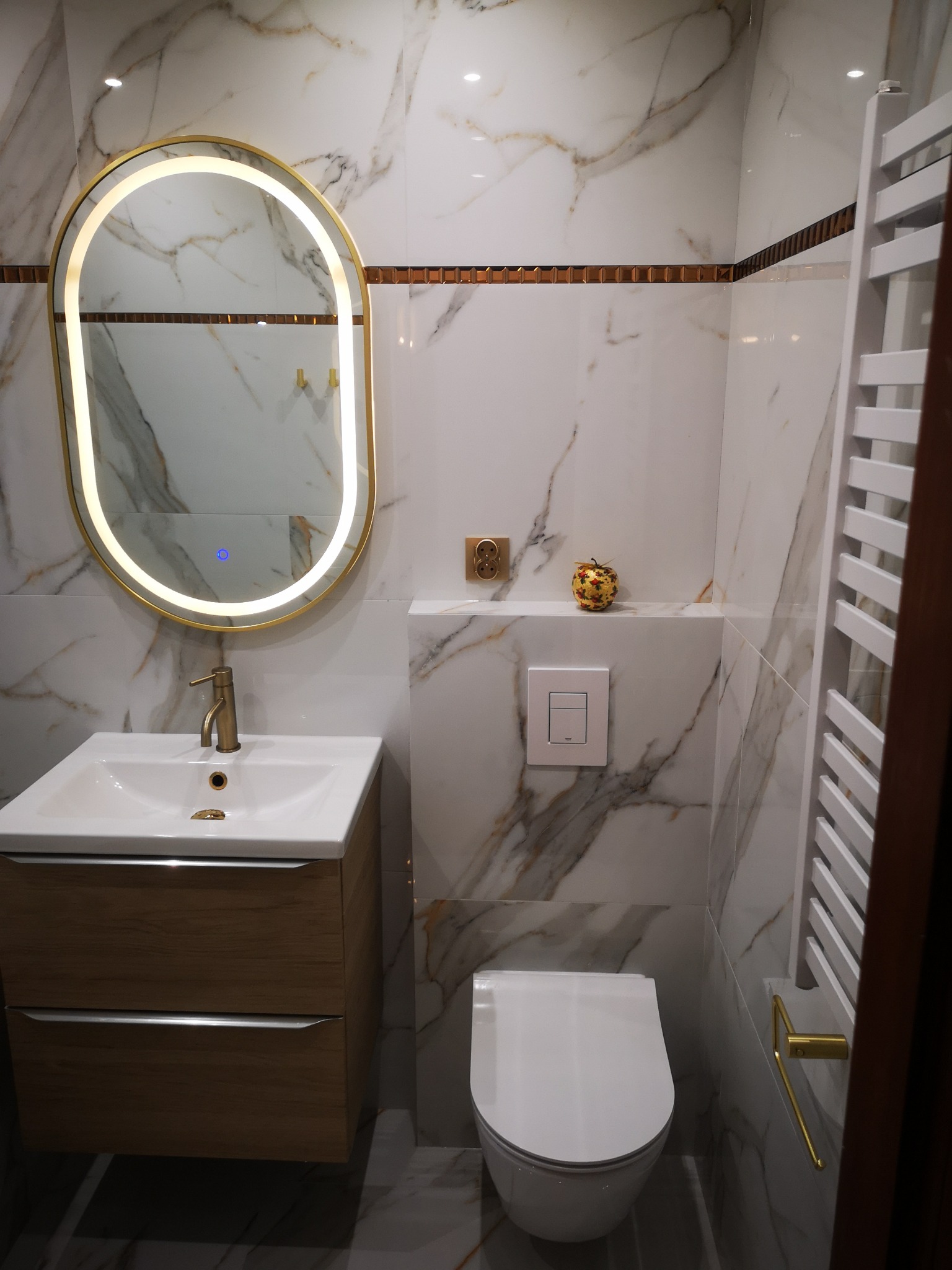 Elegancka łazienka z marmurowymi płytkami, podświetlanym lustrem, drewnianą szafką pod umywalką i grzejnikiem drabinkowym. Wykończenie w odcieniach bieli, złota i brązu.