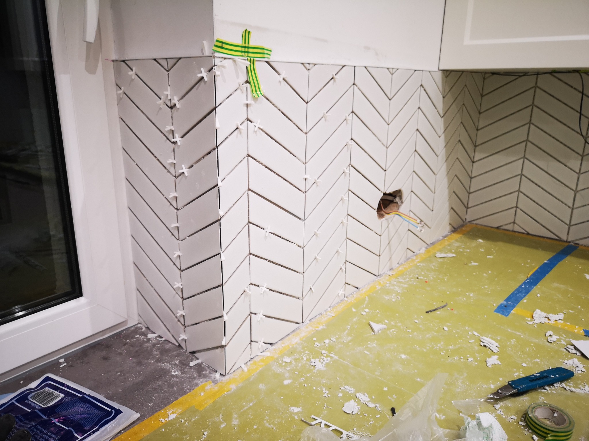 Układanie białych, podłużnych płytek w jodełkę na ścianie, widoczne krzyżyki dystansowe i taśma maskująca. Prace glazurnicze w trakcie wykańczania ściany.