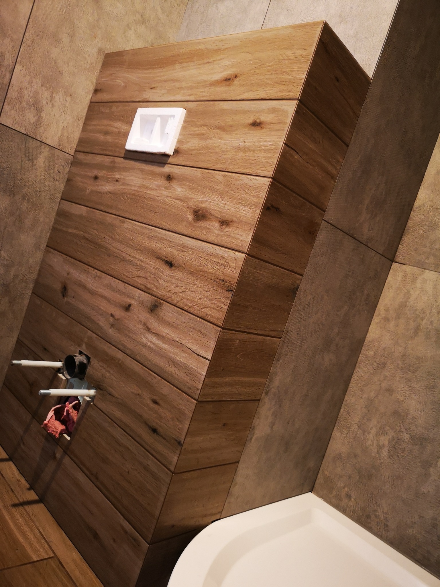 Nowoczesna zabudowa WC z drewnopodobnych płytek w łazience, widoczne przyłącza wodne i fragment wanny. Nietypowy układ płytek na ścianie.