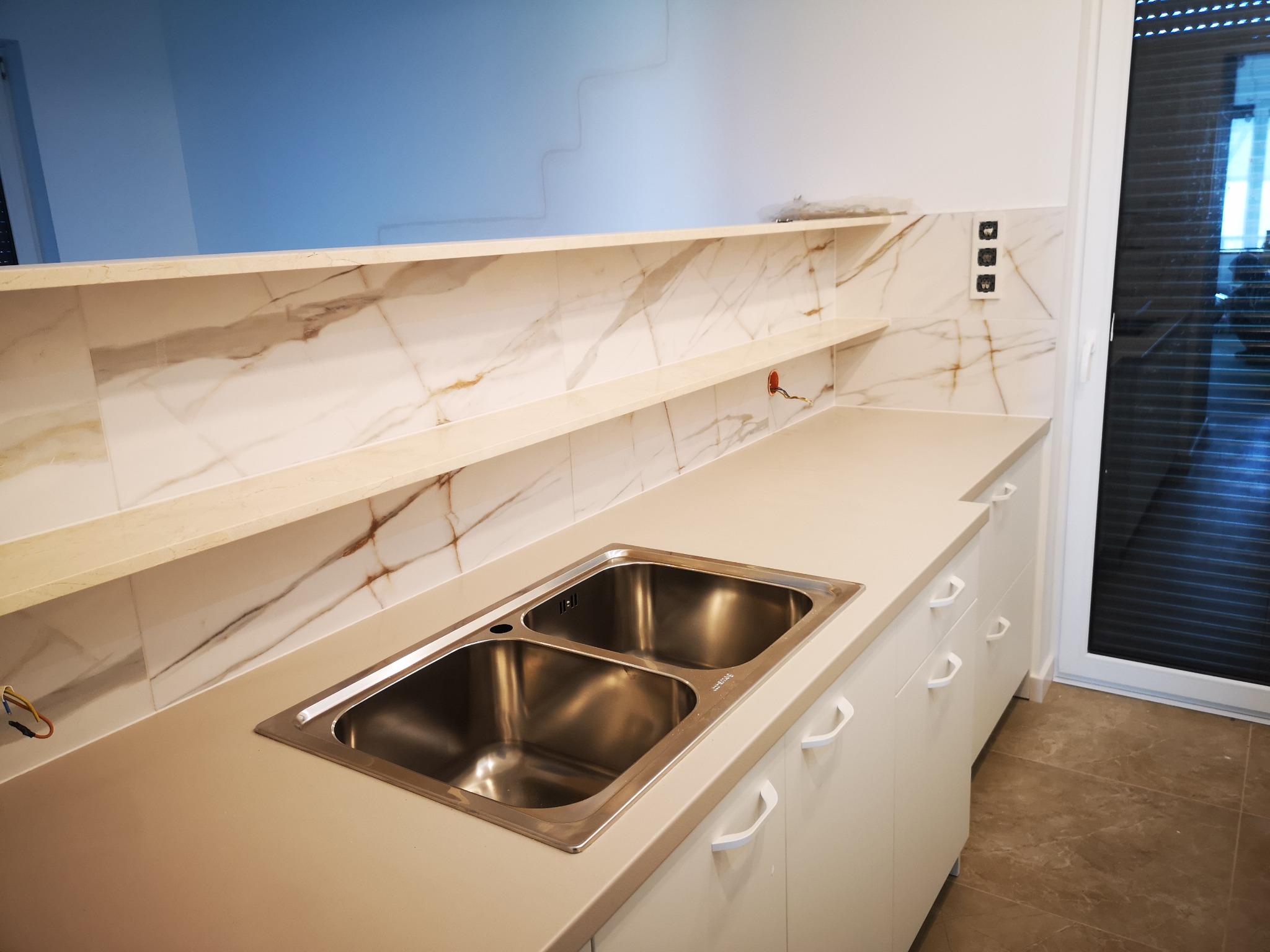 Nowoczesna kuchnia z marmurowym backsplashiem, jasnymi szafkami i dwukomorowym zlewem ze stali nierdzewnej. Widoczne gniazdka elektryczne i fragment okna.