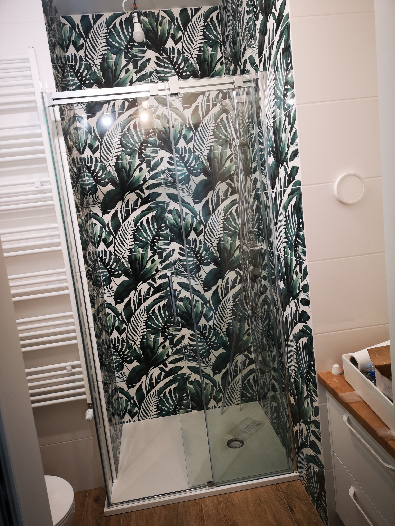 Nowoczesna kabina prysznicowa z deszczownicą, otoczona płytkami w tropikalne liście. Biały grzejnik drabinkowy i drewniany blat dodają ciepła aranżacji.