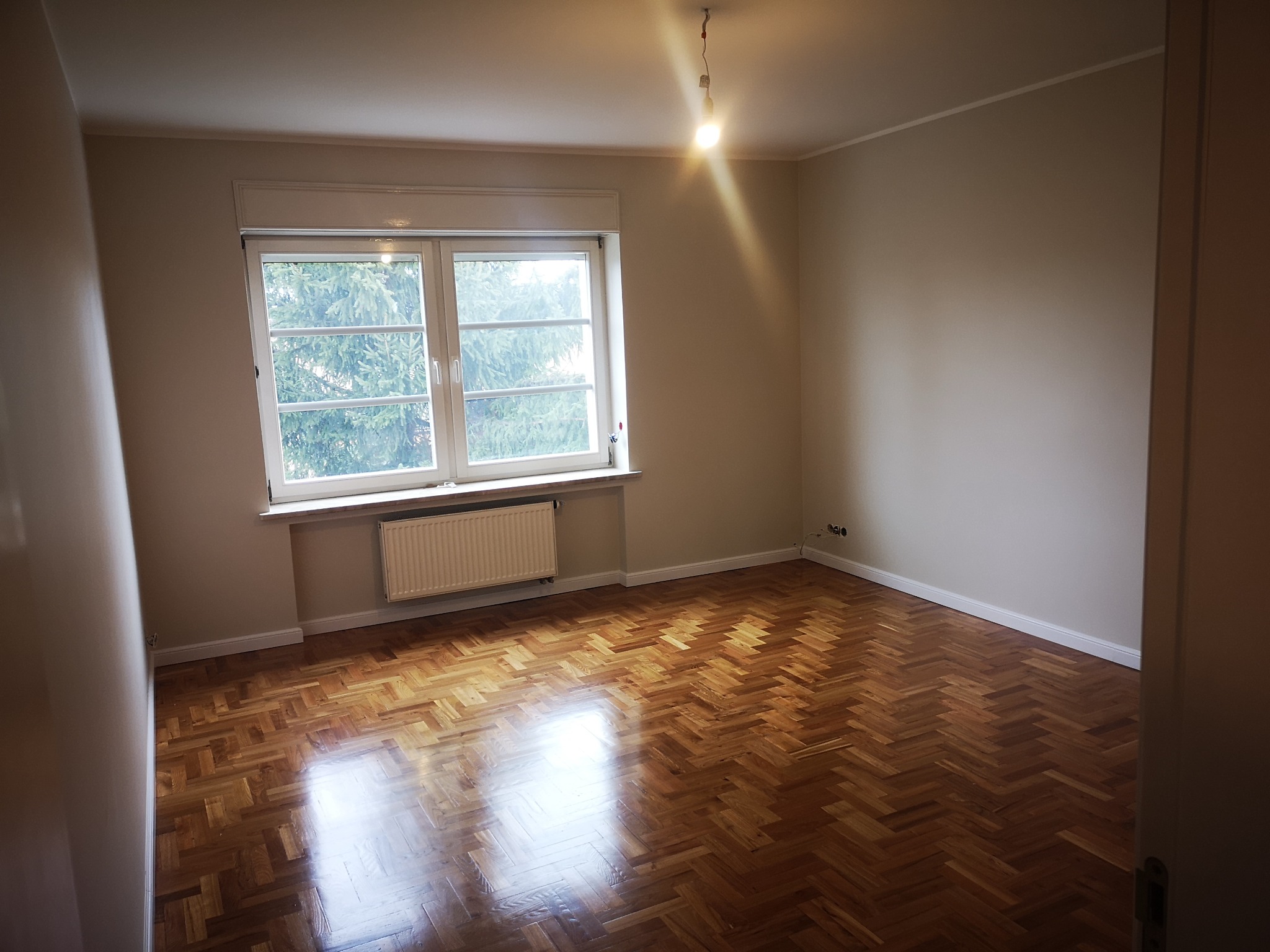 Jasne, puste wnętrze w Poznaniu z parkietem w jodełkę, białymi ścianami i oknem. Widoczny grzejnik i wisząca żarówka, podkreślające minimalizm aranżacji.