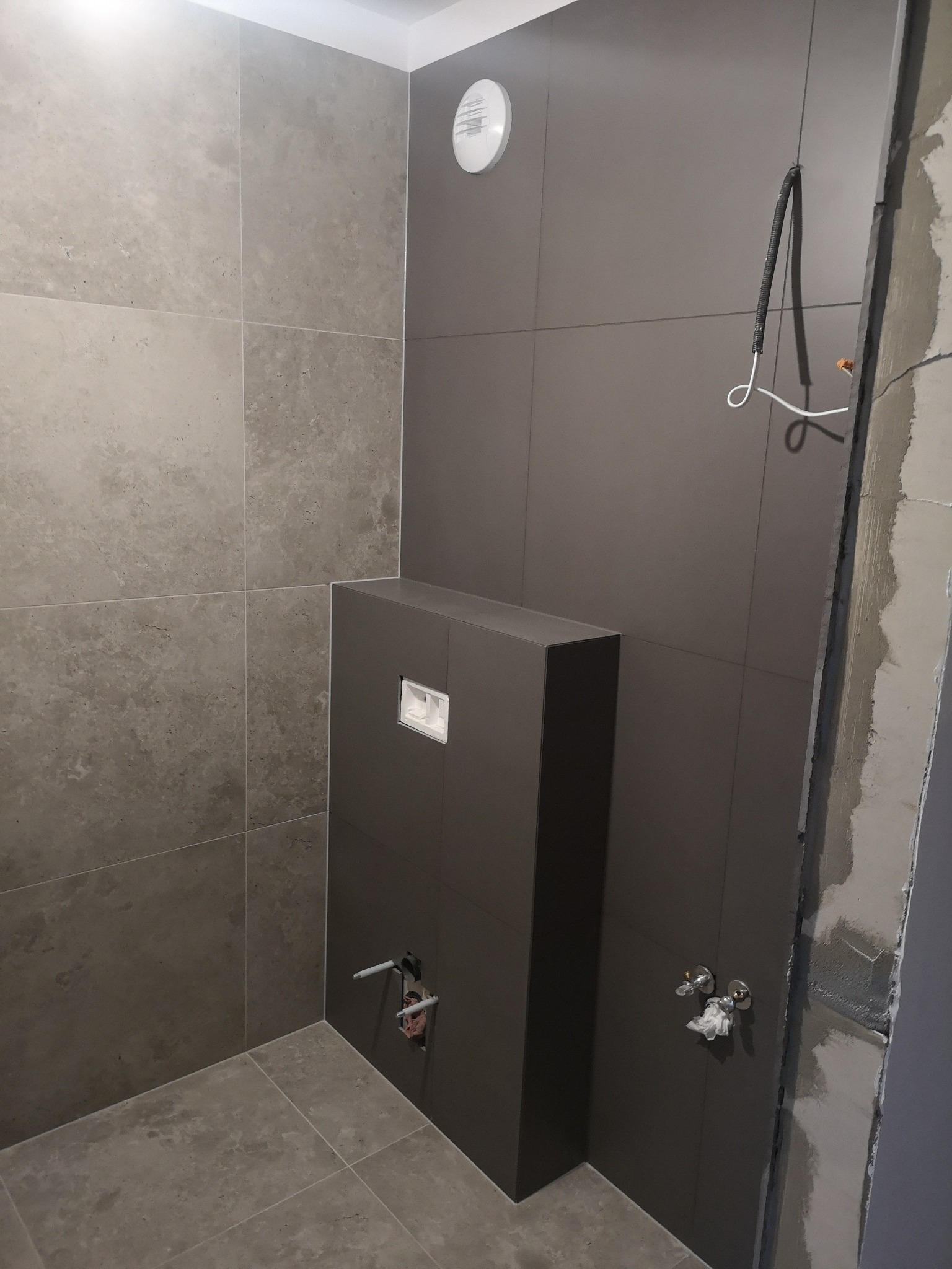 Wykończona łazienka z szarymi płytkami, zabudowanym stelażem WC i widoczną instalacją. Częściowo wykończone ściany, wentylator sufitowy.