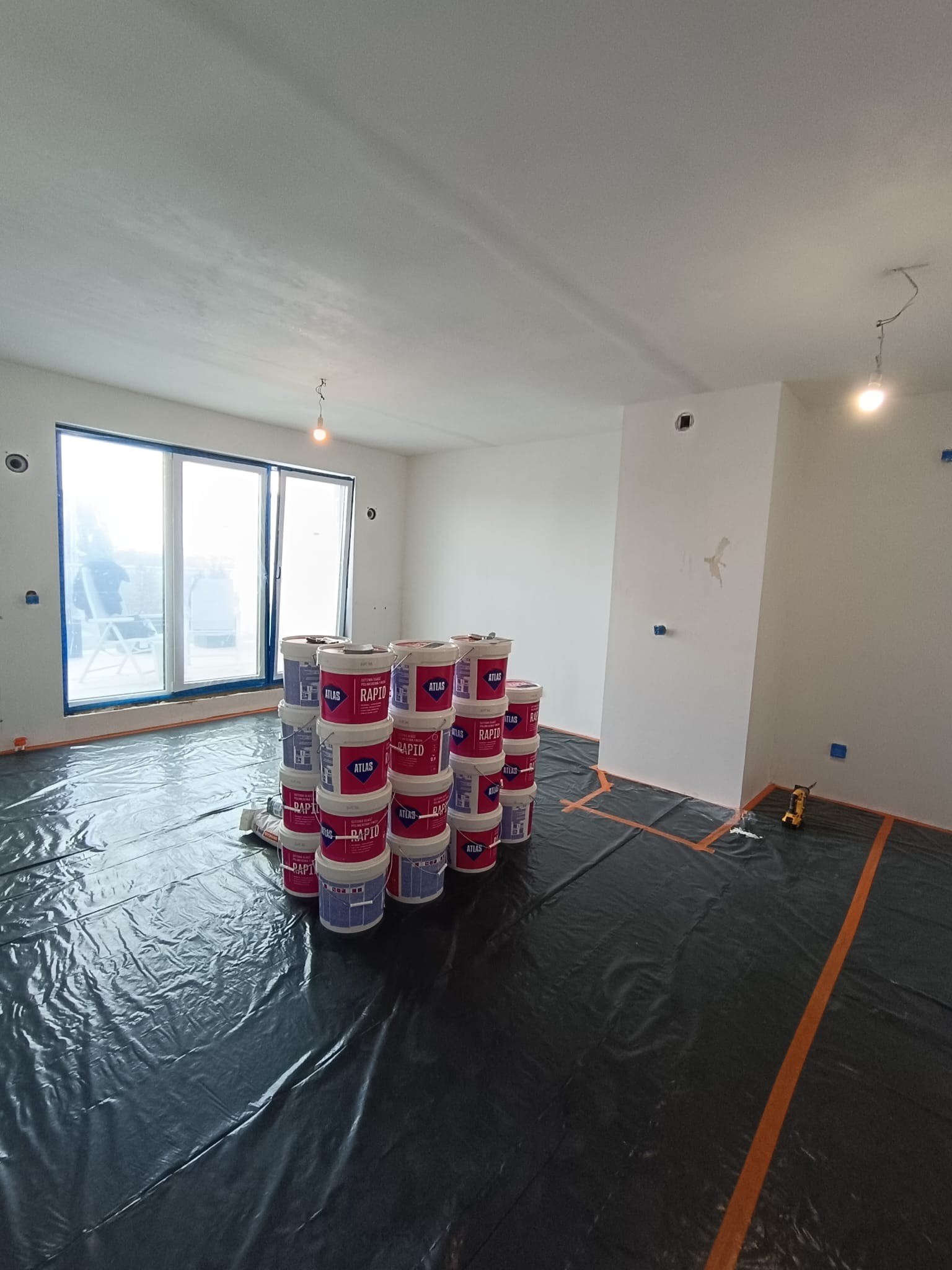 Przygotowania do malowania: pokój zabezpieczony folią, ustawione wiadra z farbą Atlas Rapid. Widoczne gładkie ściany, okno z niebieską taśmą malarską i narzędzia.