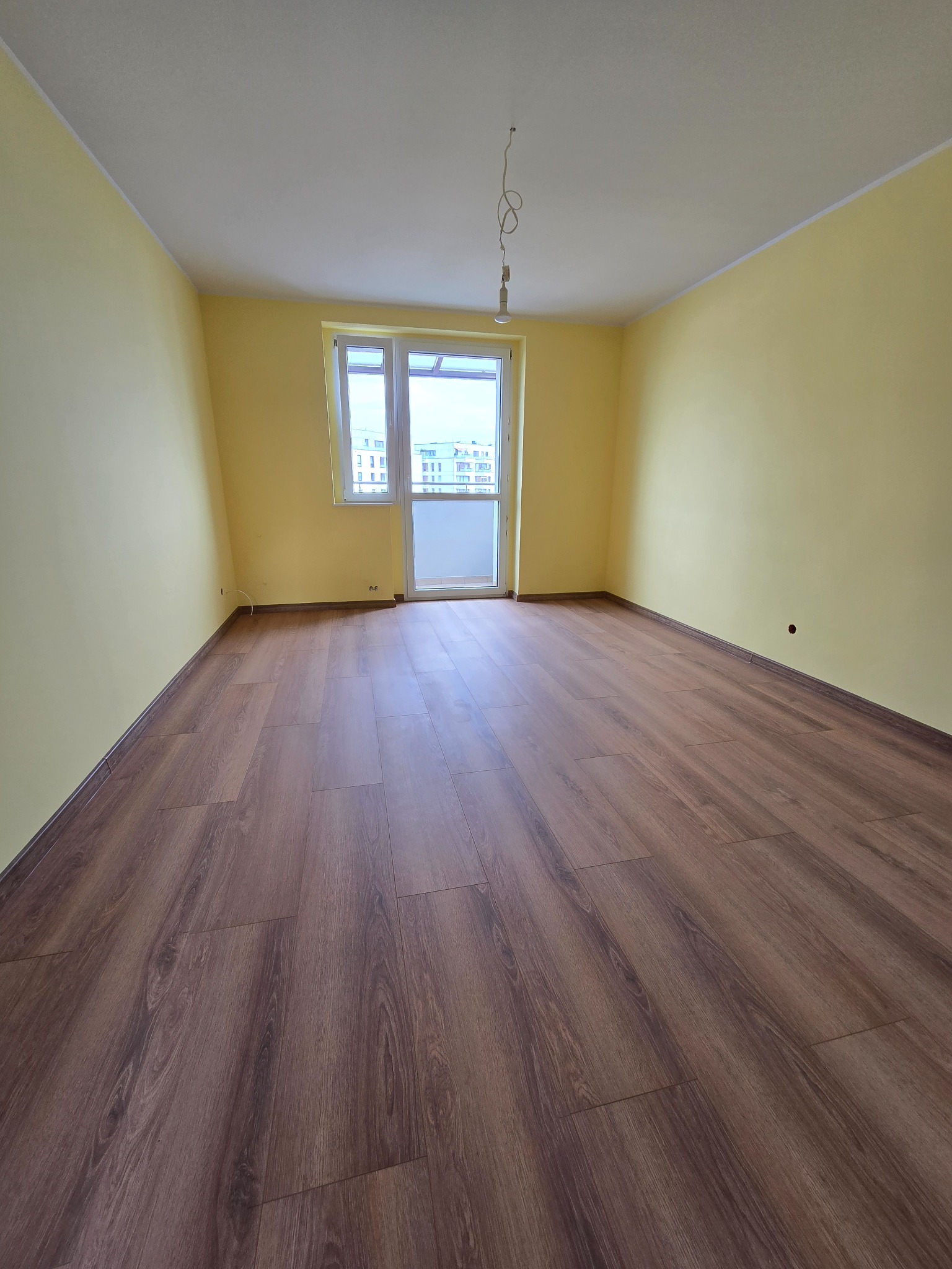 Pusty pokój z panelami podłogowymi w odcieniach brązu i żółtymi ścianami. Widoczne okno balkonowe i sufit z wiszącym przewodem. Podłoga ułożona wzdłuż dłuższego boku pokoju.