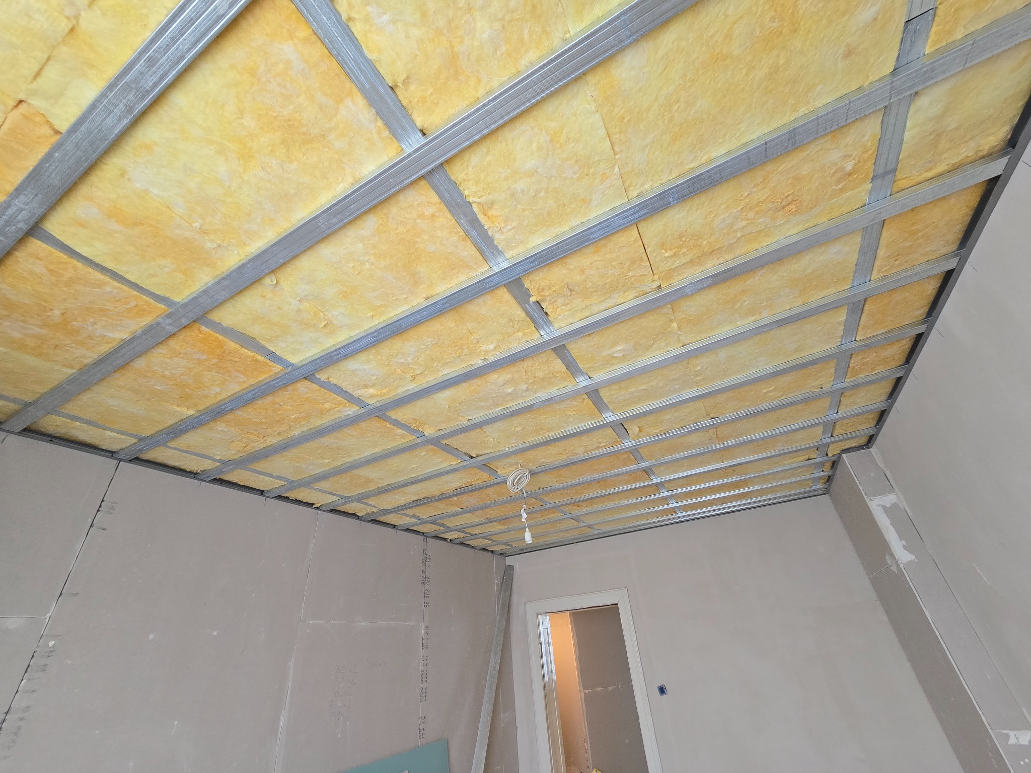 Montaż sufitu podwieszanego: metalowa konstrukcja z izolacją z wełny mineralnej, widoczne płyty gipsowo-kartonowe na ścianach, przygotowanie do dalszych prac wykończeniowych.